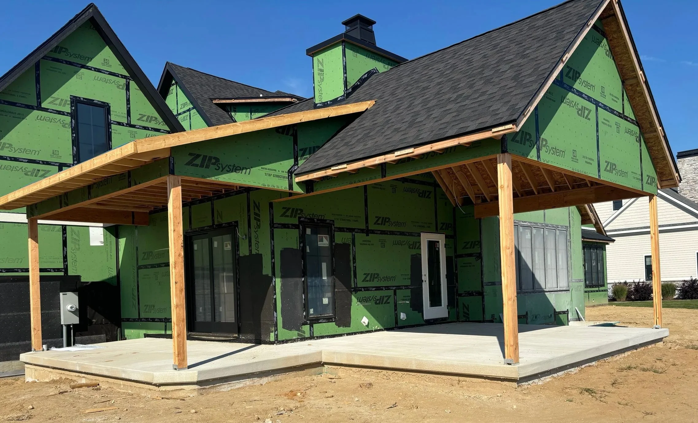 A poured concrete back patio and covered porch framing take shape on a custom home in Northwest Indiana, highlighting the installation of green sheathing panels and taped seams for weatherproofing.