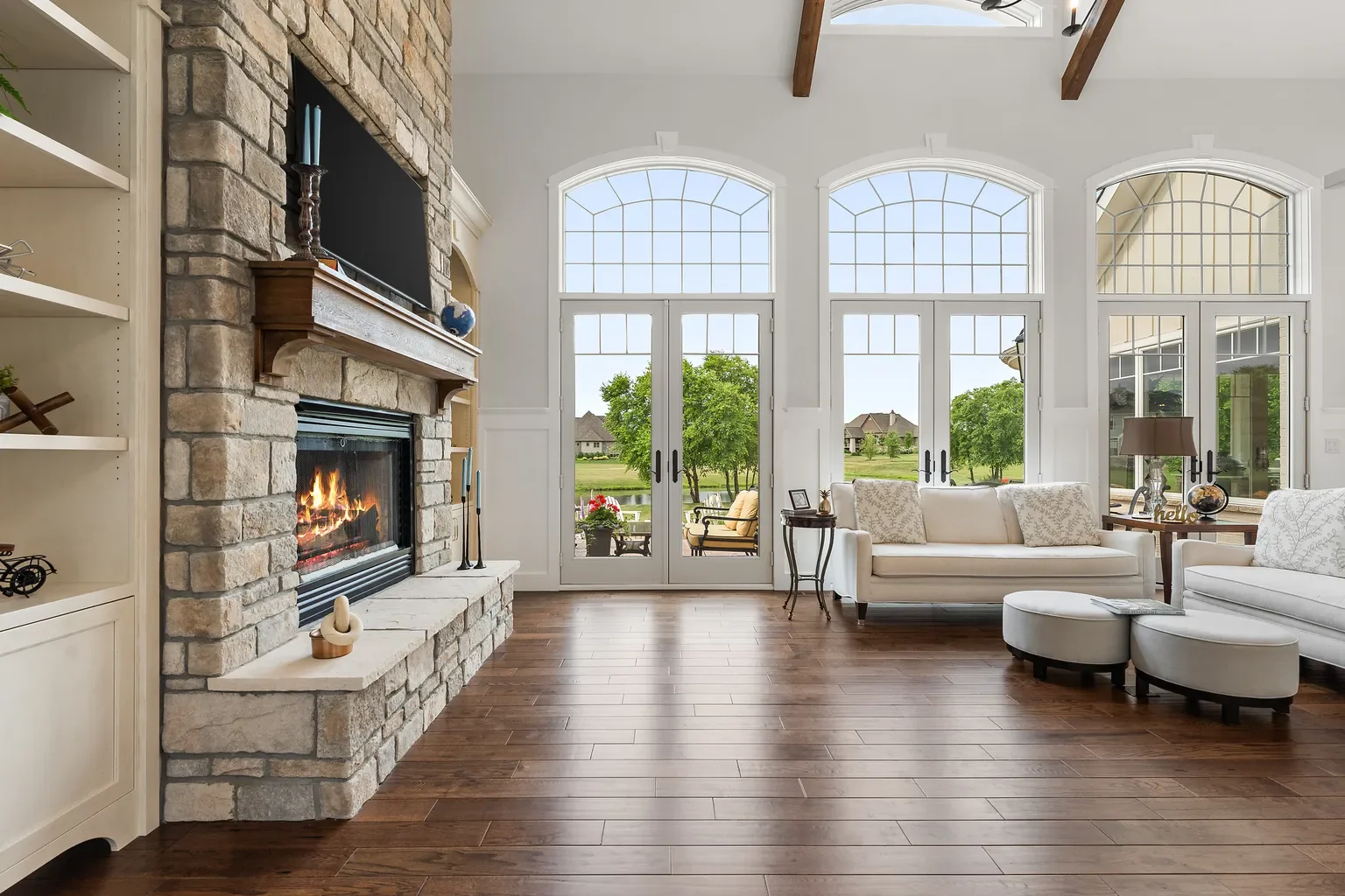 Crafted in Sand Creek, a welcoming living space highlights a floor-to-ceiling stone fireplace alongside a trio of large arched patio doors that invite natural light across the hardwood flooring.