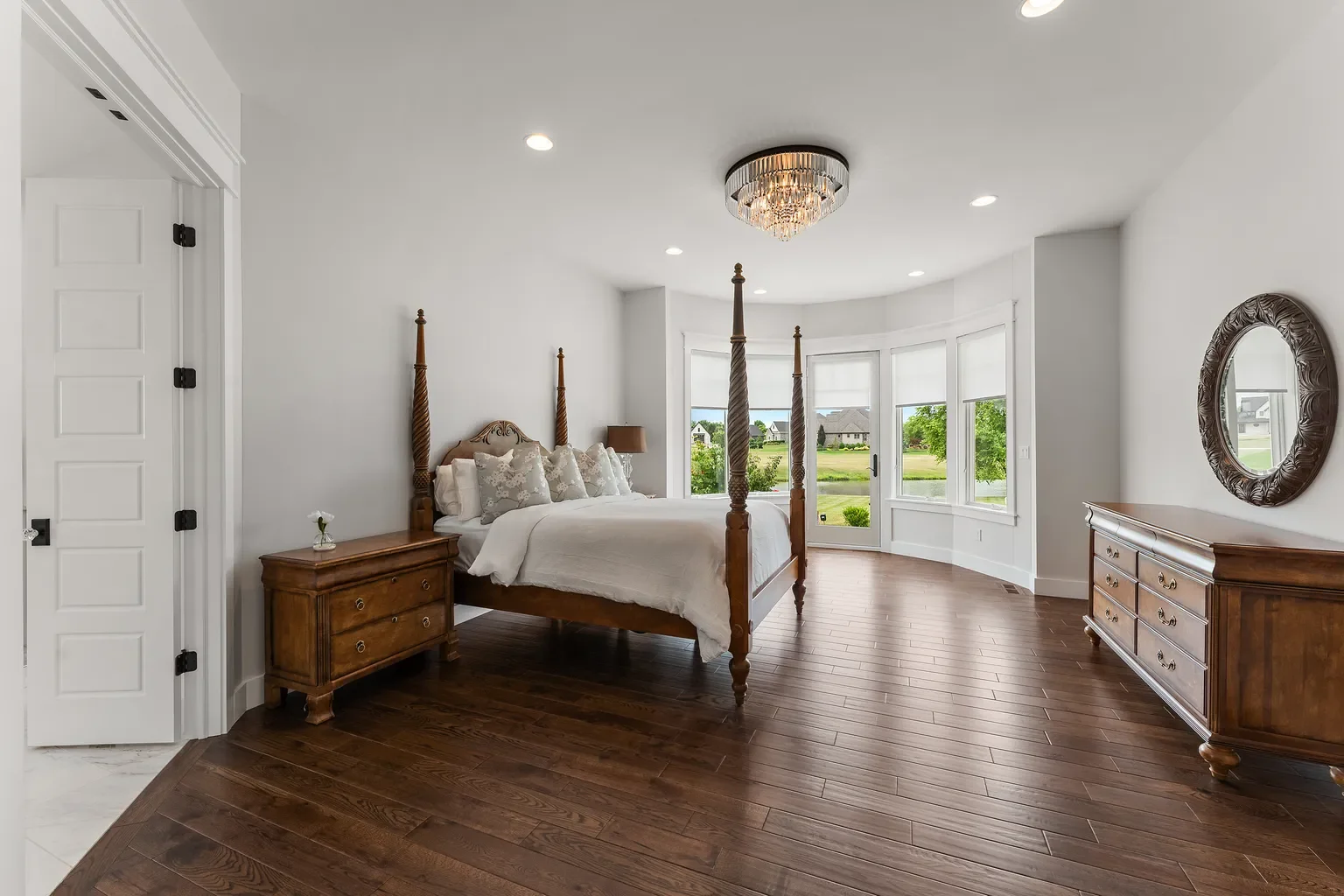 A bright bedroom constructed by Booras Builders in Northwest Indiana featuring a classic bay window alcove, rich hardwood floors, crisp white crown molding, and recessed lighting. The bedroom has a dresser, nightstand, and bed with a wooden frame.