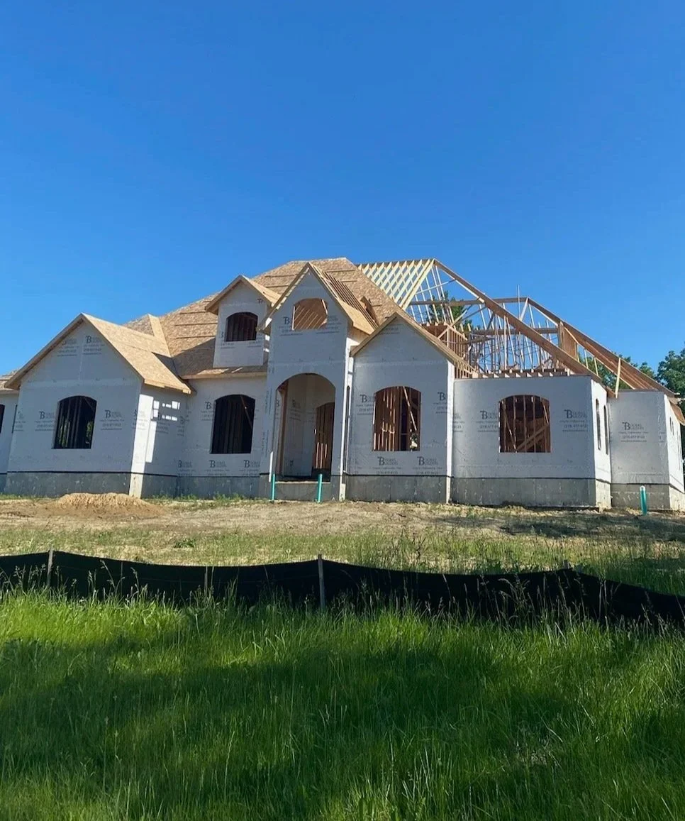 Under a clear blue sky, the roof truss installation phase progresses on a sprawling estate, highlighting precision wood roof framing, arched entryway details, and protective house wrap on the exterior walls.
