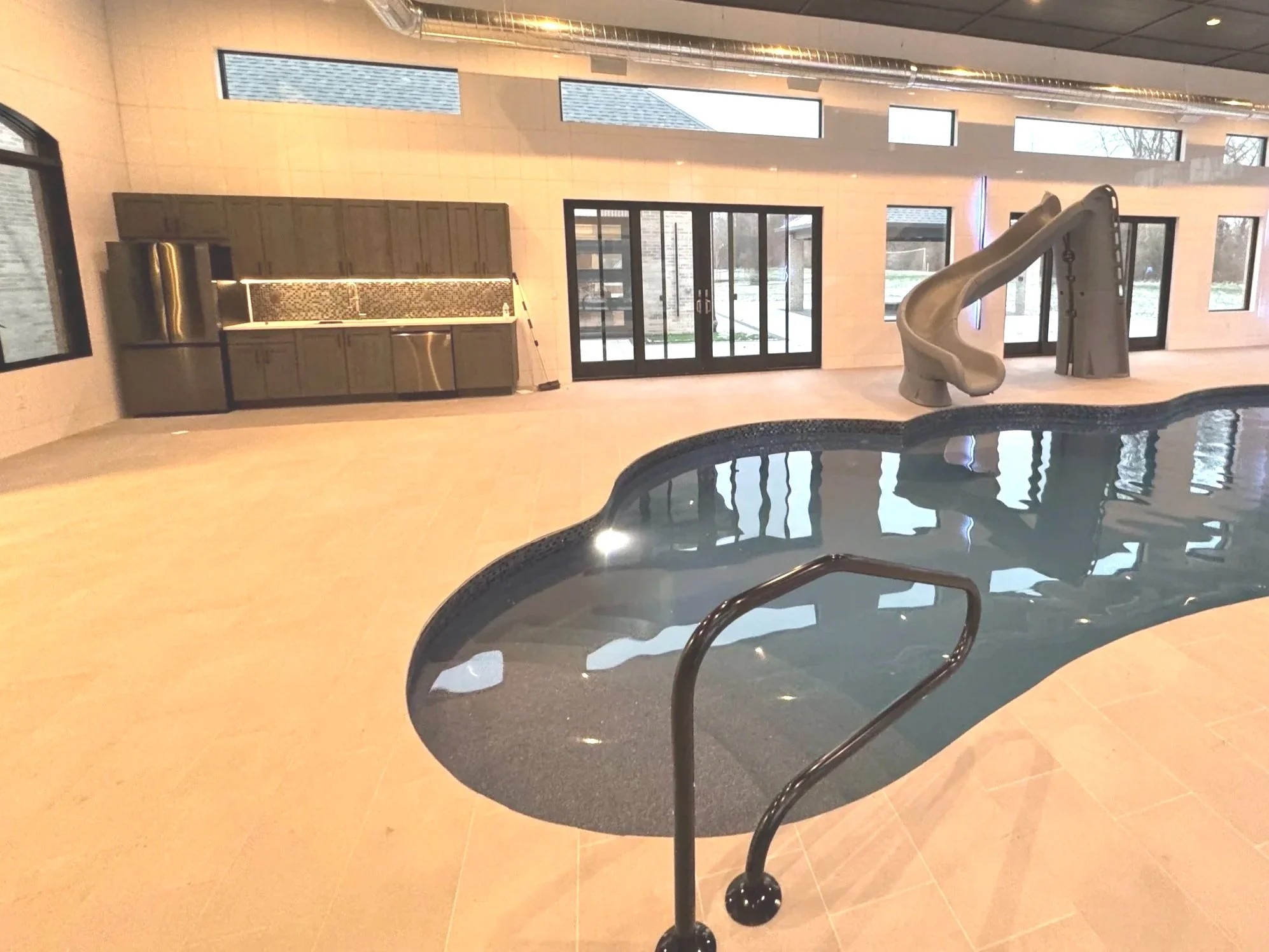 An indoor swimming pool house built by Booras Builders in Chesterton features a custom pool with a water slide, built-in kitchenette, and large multi-panel glass patio doors surrounded by tile walls and transom windows.