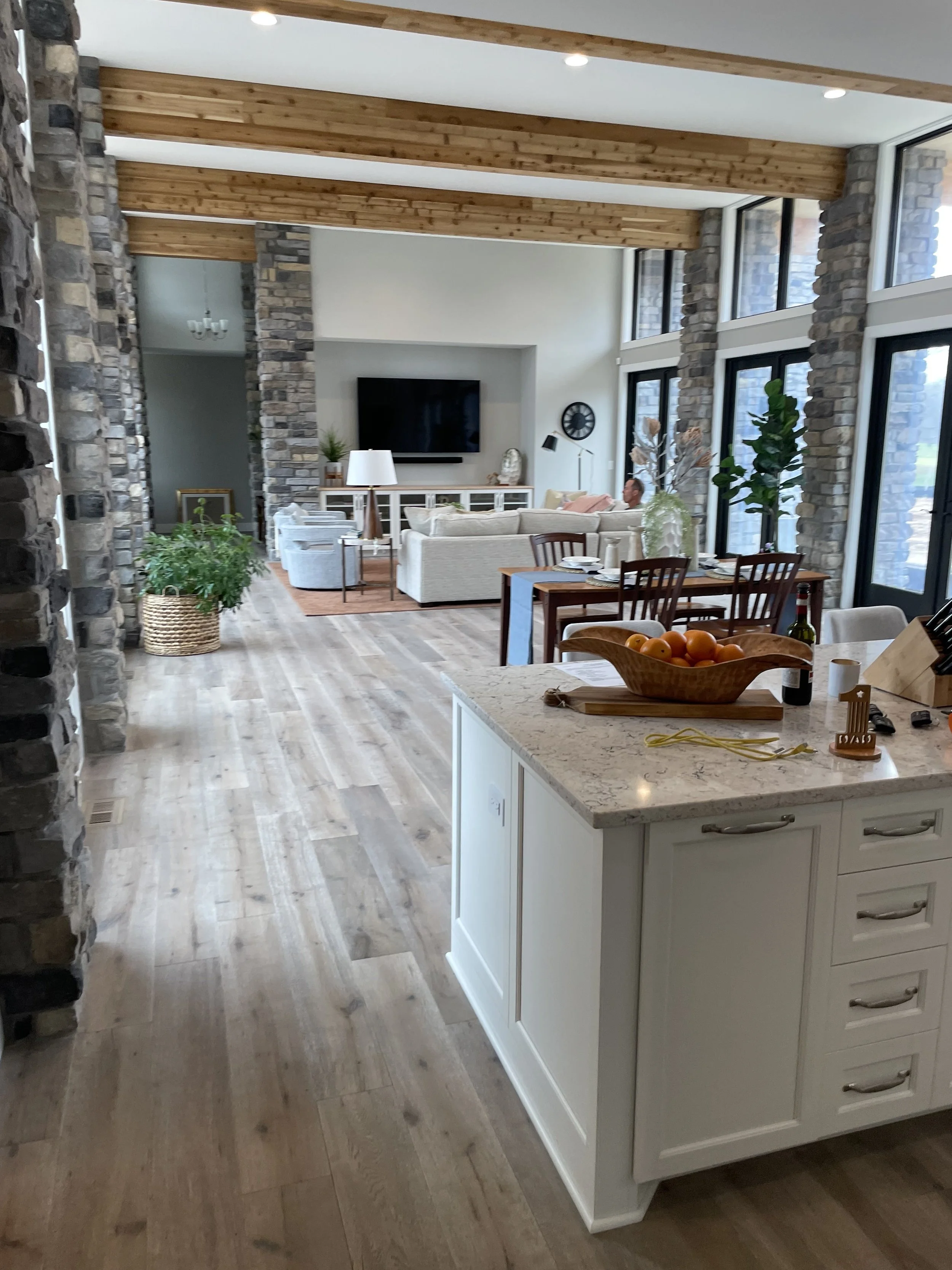 Exposed timber ceiling beams and floor-to-ceiling stone pillars line the open-concept floor plan of this residential construction project built in Chesterton, connecting a white kitchen island to a sunlit living space.
