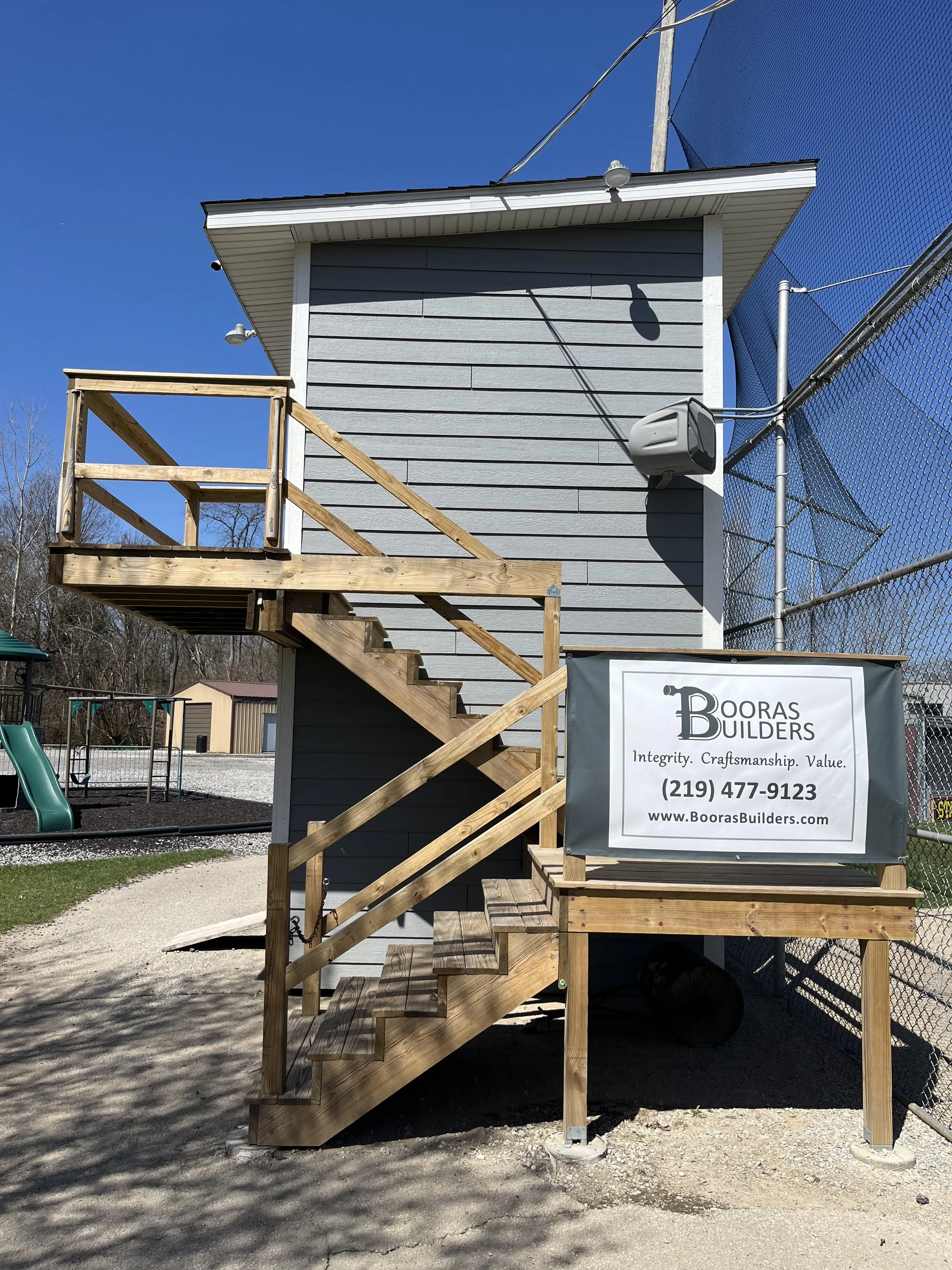 A pressbox at State Park Little League in Porter, Indiana, with gray siding and a wooden staircase situated behind a chain link fence and netting to protect it from foul balls. The building was custom-built and donated by Booras Builders.