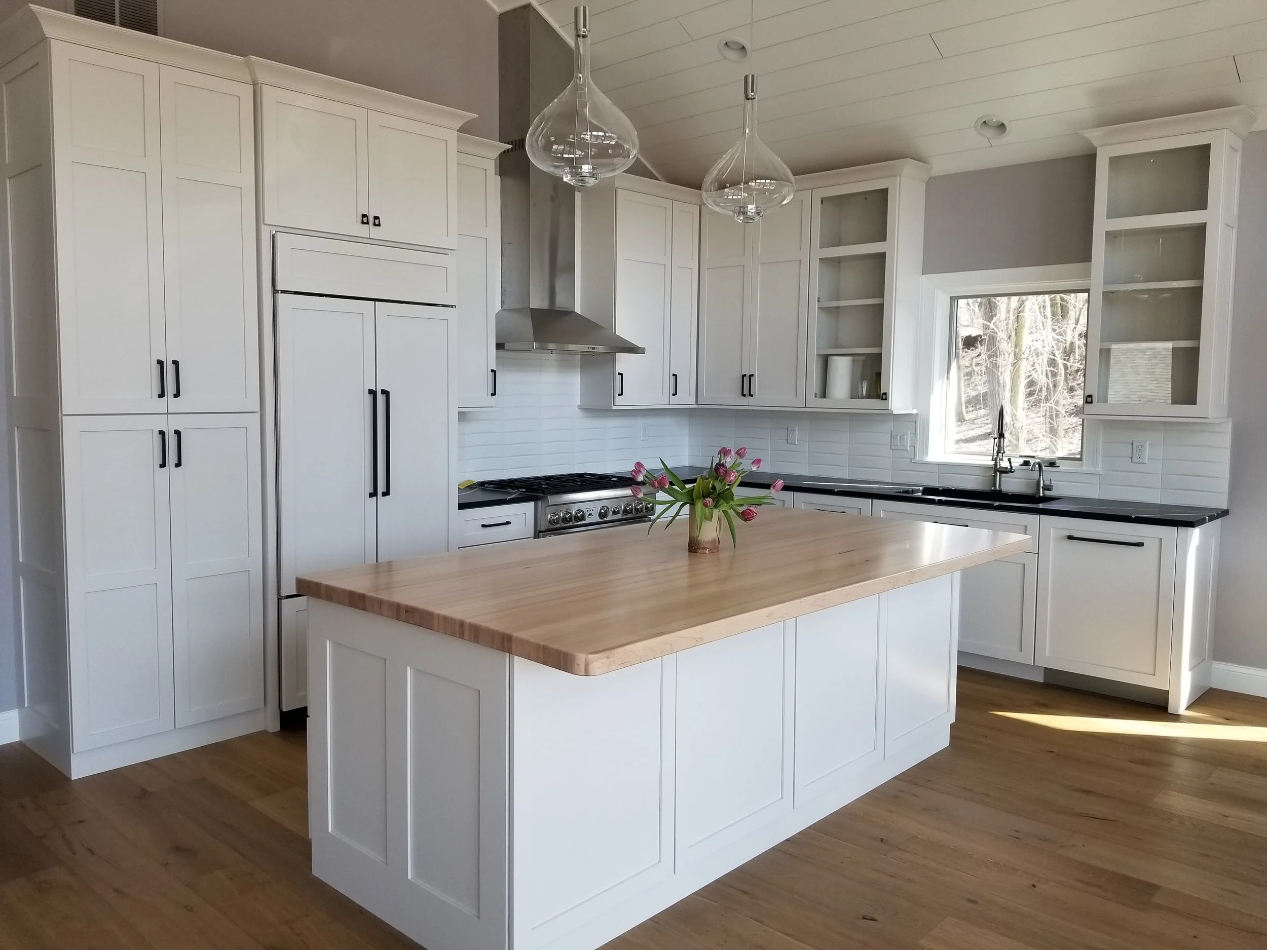 Contemporary residential construction by Booras Builders in Porter County showcases a bright kitchen centered around a thick butcher block island countertop, clear teardrop pendant lights, hardware flooring, and a classic shiplap ceiling.