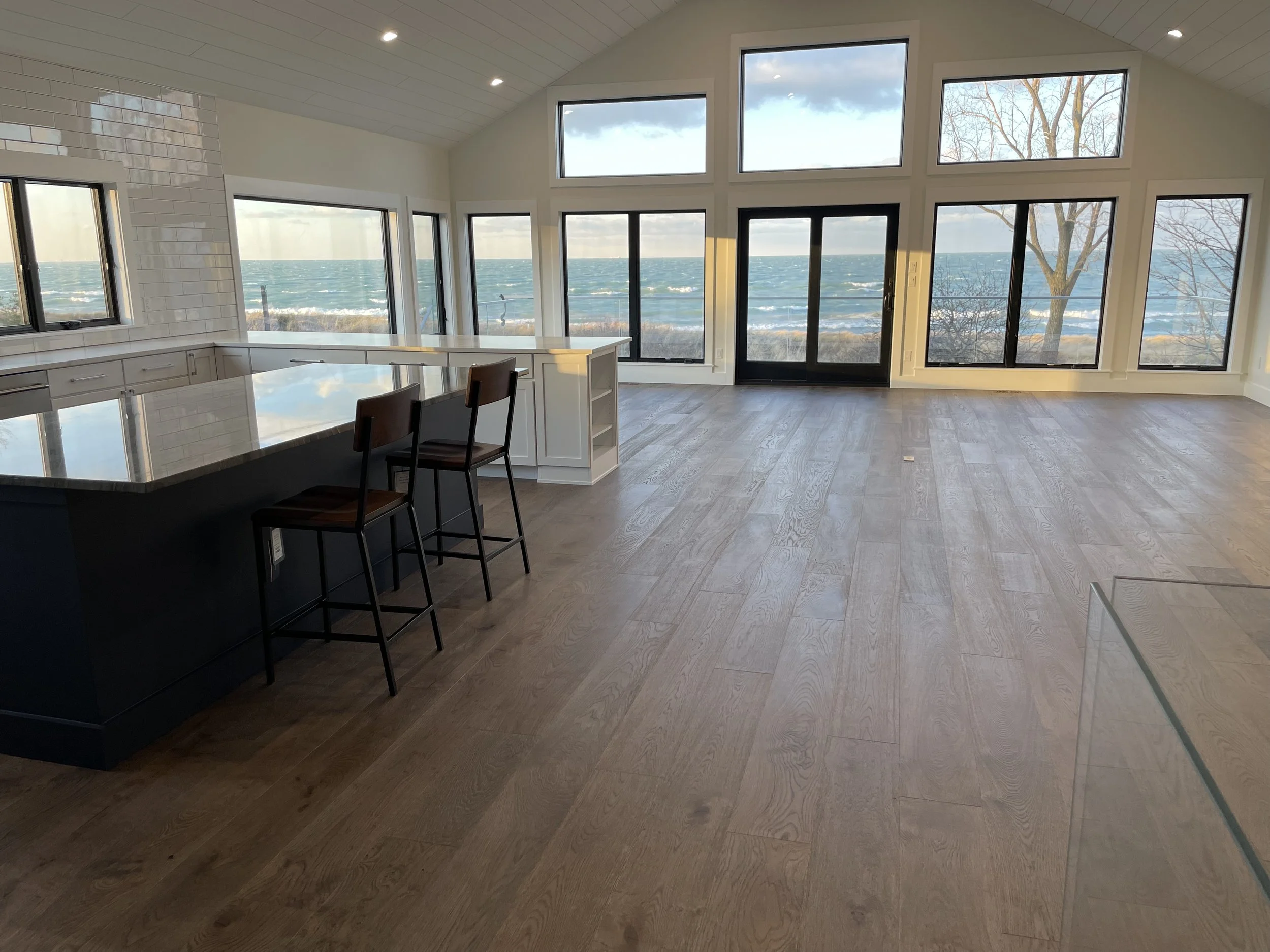 In a bespoke lake house built by Booras Builders, the waterfront great room and kitchen pair wide-plank wood flooring with a white shiplap vaulted ceiling, framed by an expansive wall of windows that overlook the waves of Lake Michigan.