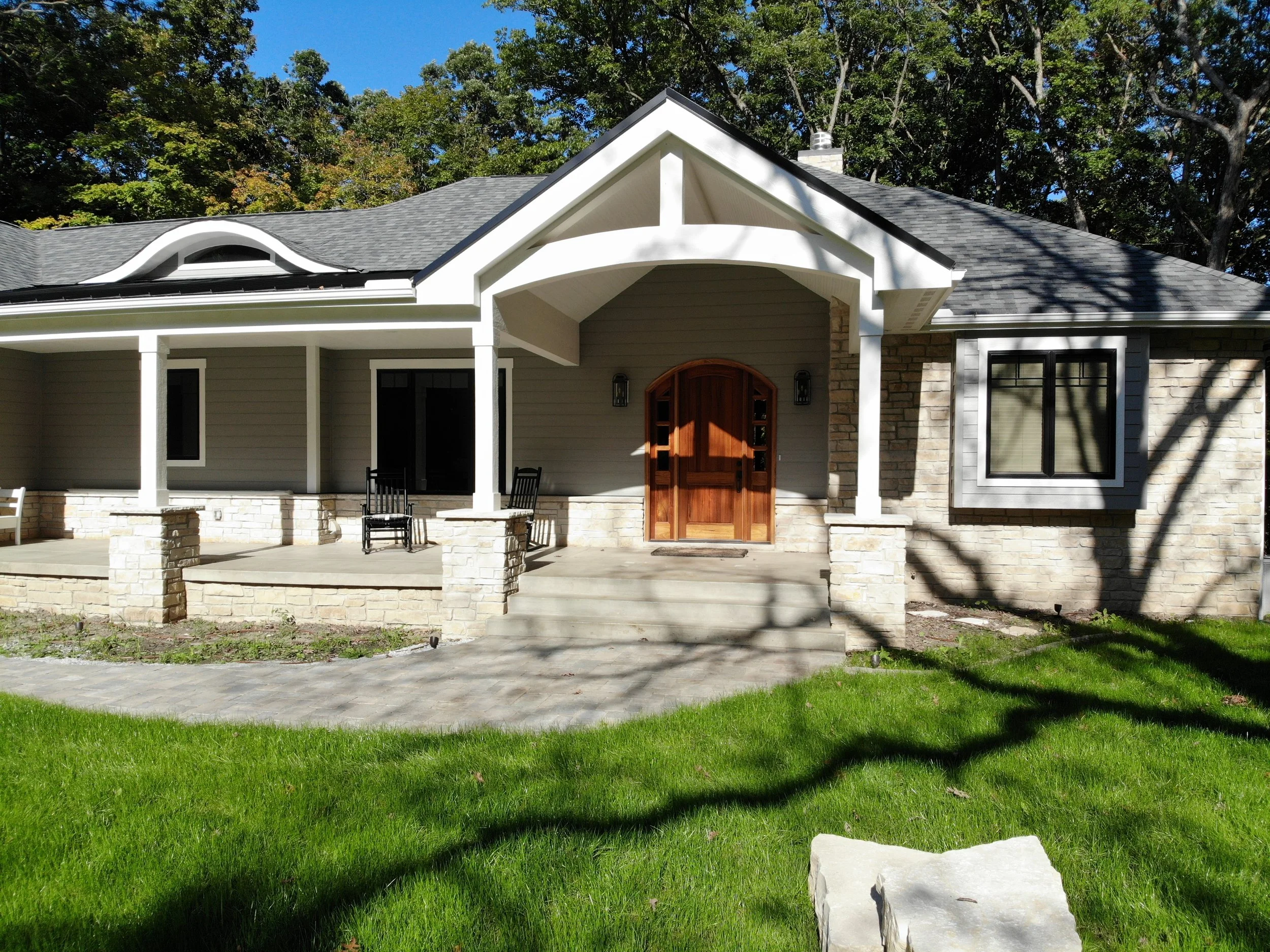 Elegant front exterior of a bespoke home in Porter County with a spacious covered porch, stone-based columns, gray horizontal siding with white trim, natural limestone veneer, black-framed windows, an arched wood front door, and a paved walkway.