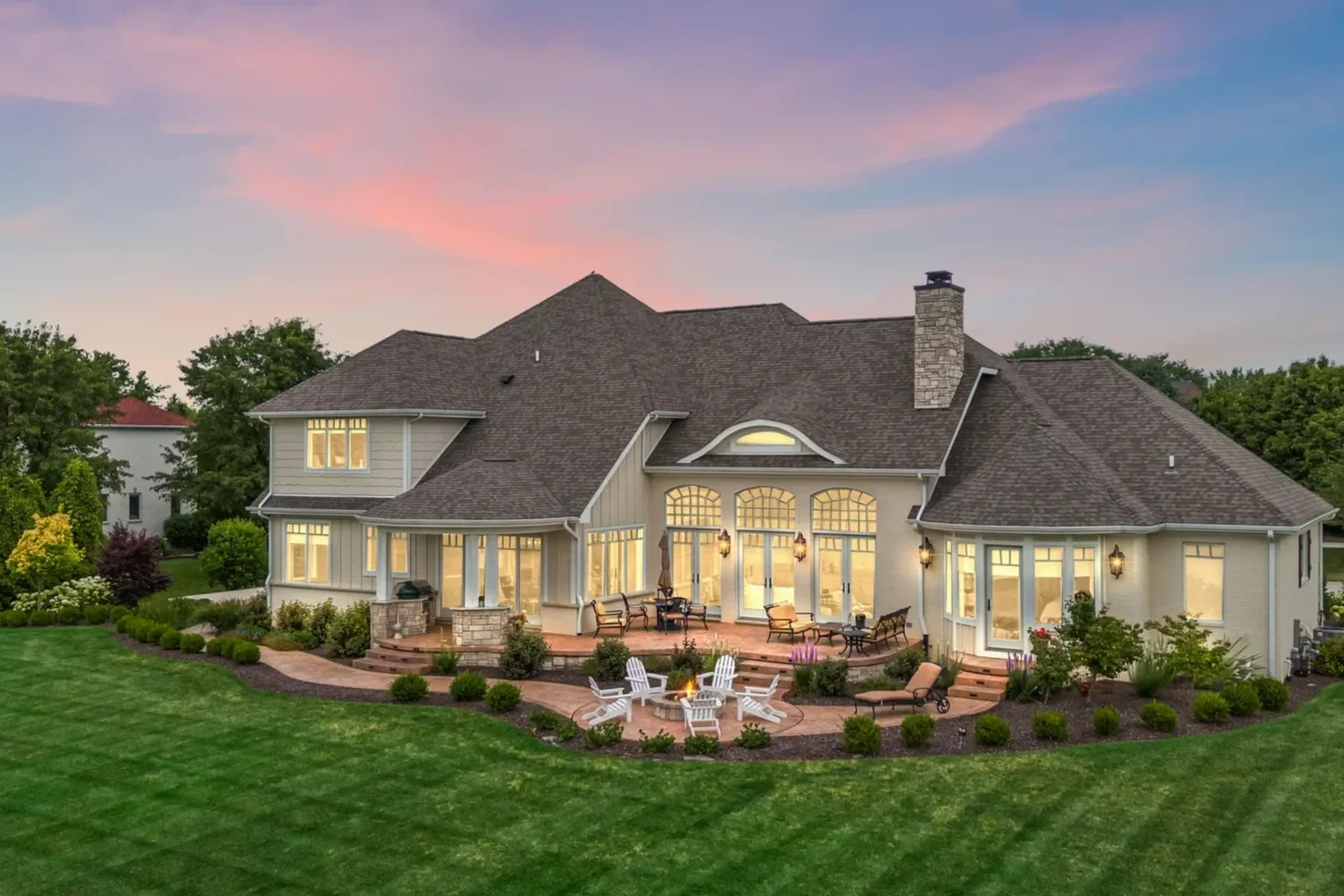 In Chesterton, a beautifully landscaped backyard features a home custom-crafted by Booras Builders. It has large panoramic windows, warm lighting, a spacious patio area, lush green landscaping, and cool roof details, including an eyebrow dormer.