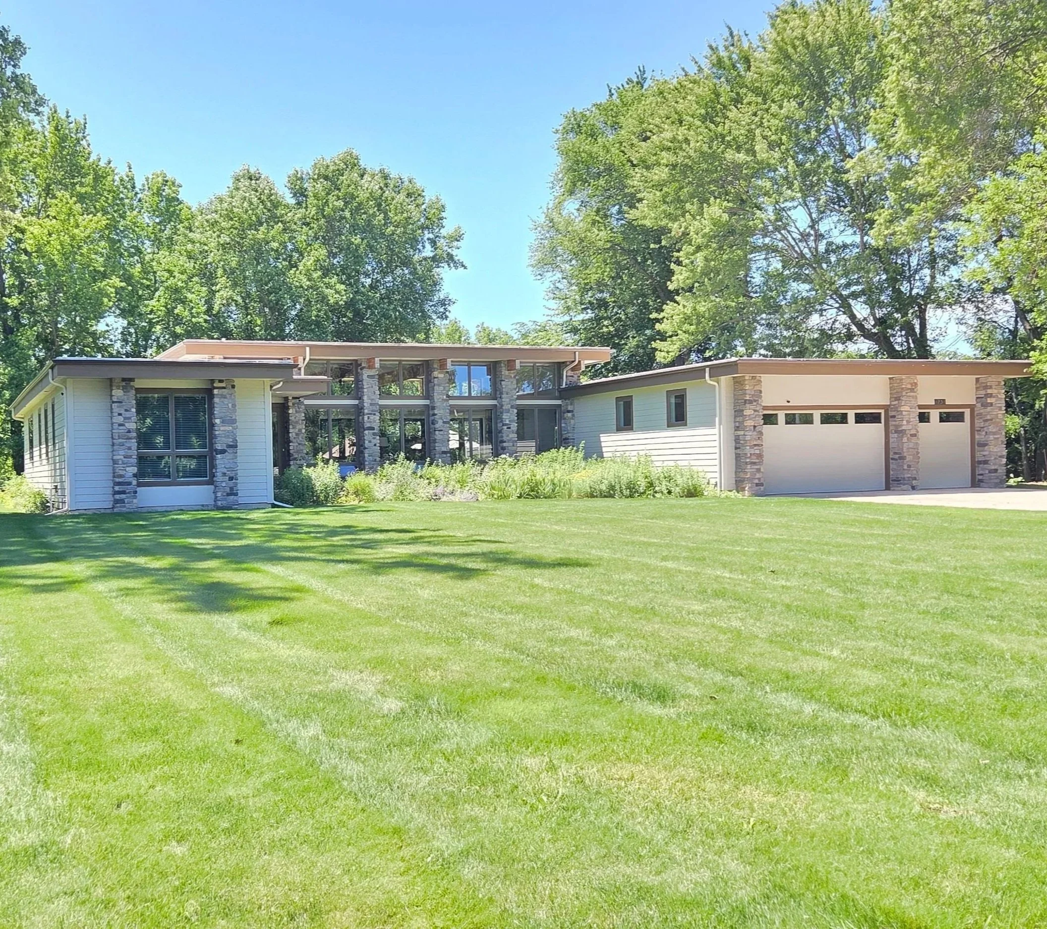 A single-story Chesterton home featuring stone pillars, large glass panels, and an attached garage. A massive, perfectly manicured green lawn dominates the foreground, framed by lush trees and a partly cloudy sky.