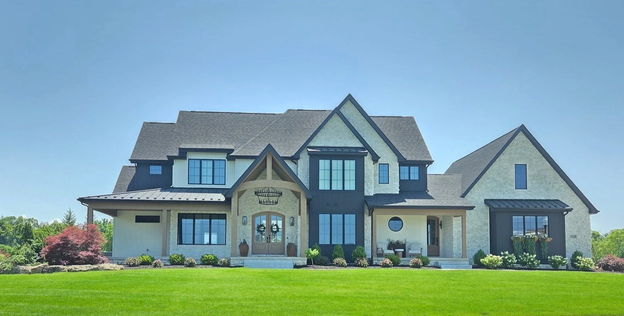 This luxury Booras Builders estate exhibits intricate multi-gabled rooflines, seamlessly integrated white masonry, contrasting black board-and-batten extensions, heavy timber-framed structural vaults, and a distinctive porthole accent window.