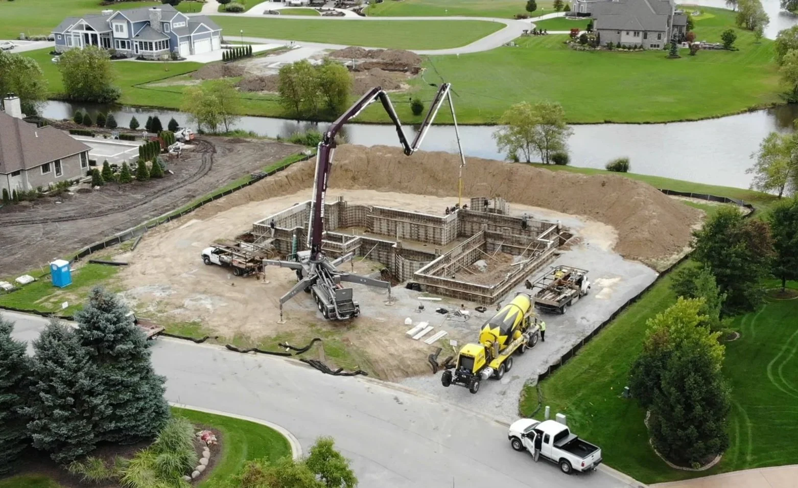 A construction site in Chesterton where a house foundation is being built by Booras Builders, with concrete being poured by a large yellow cement mixer truck and a concrete pump.