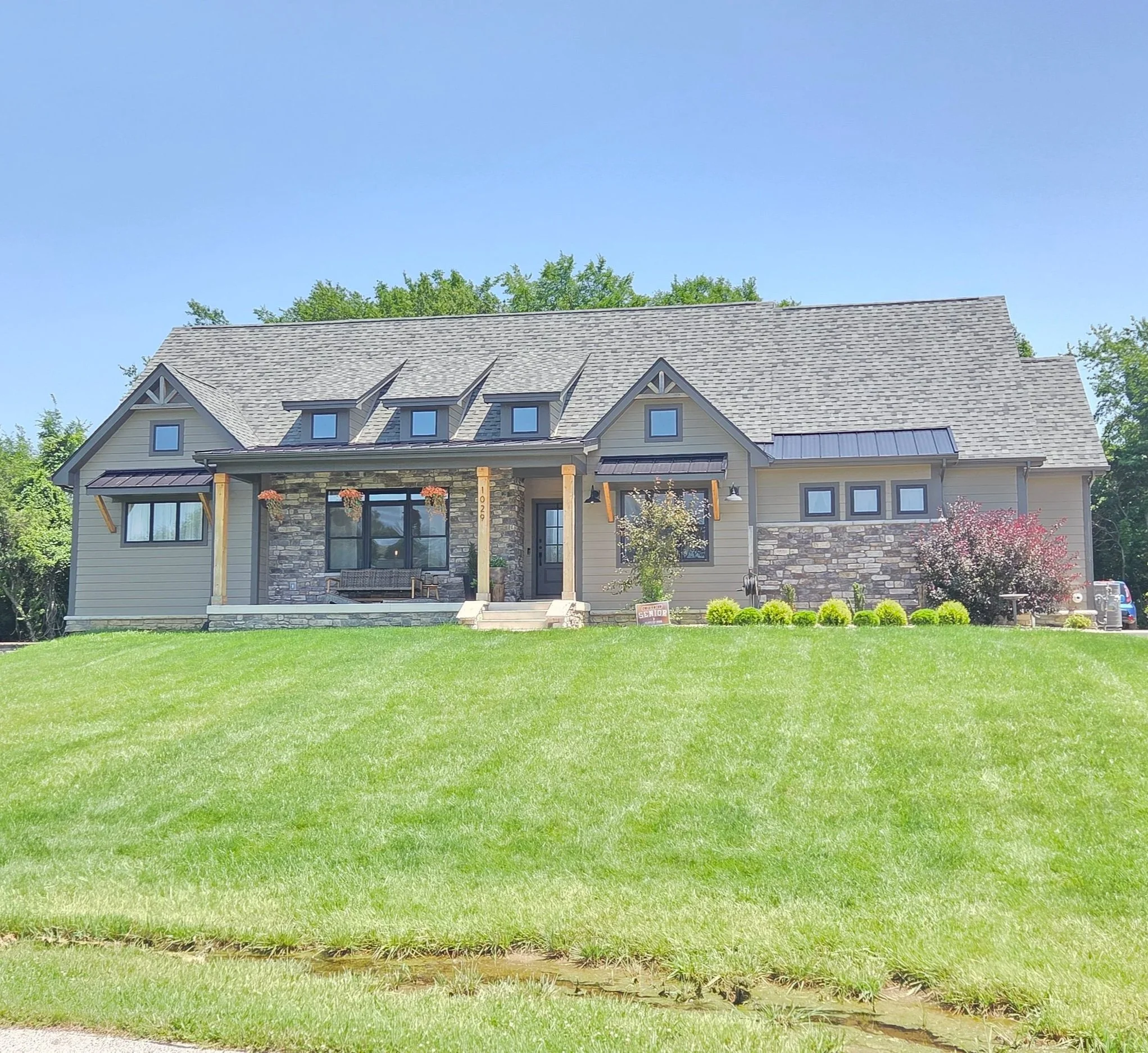 This modern custom built home features a wide covered front porch, three distinct shed dormers, stone wainscoting accents, dark window trim, and a manicured sloping lawn.