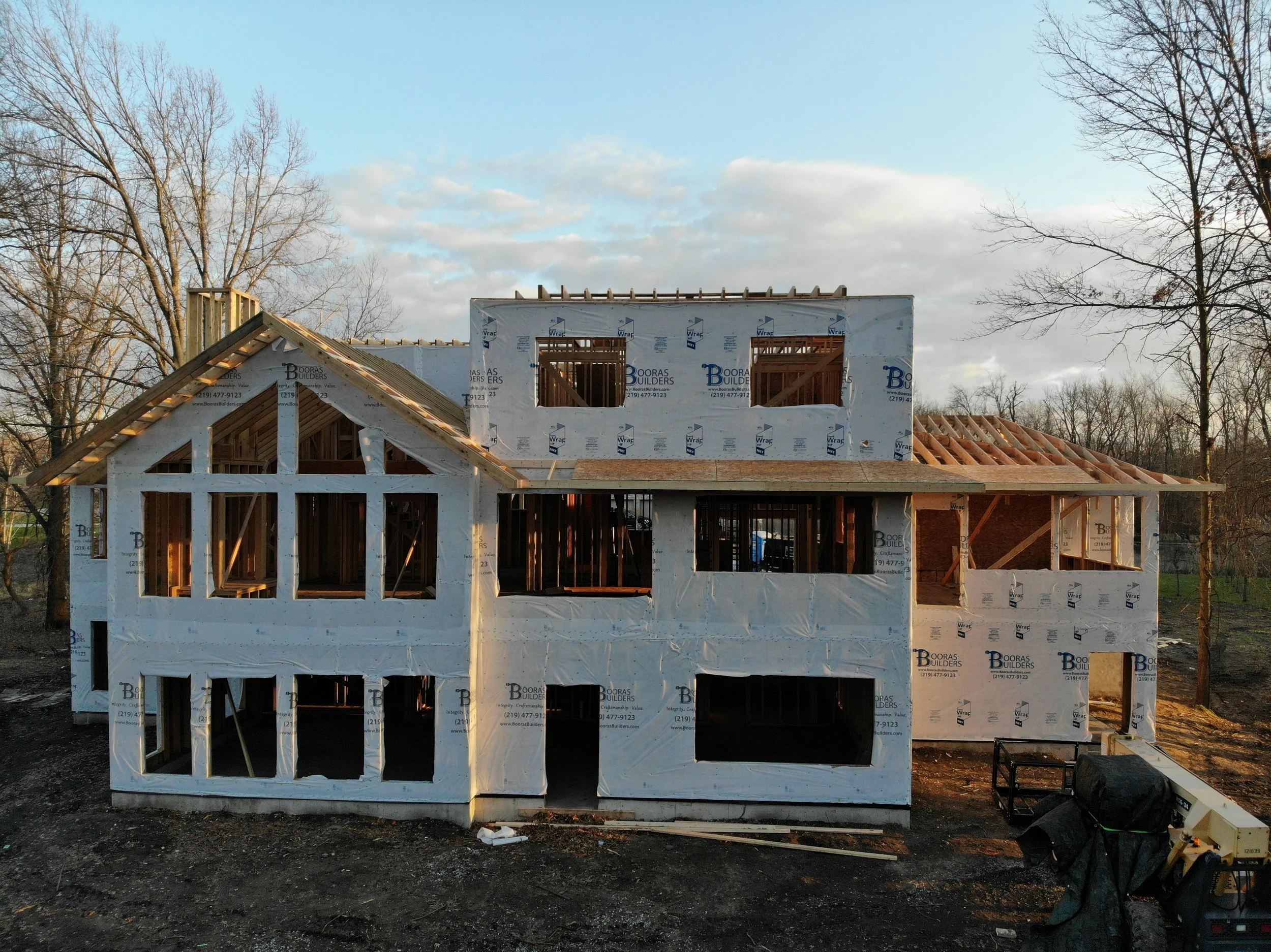 The wood framing phase of a multi-story custom home being built in Chesterton by Booras Builders showcases the exterior house wrap, precise window cutouts for a great room, and roof rafters under a partly cloudy sky.