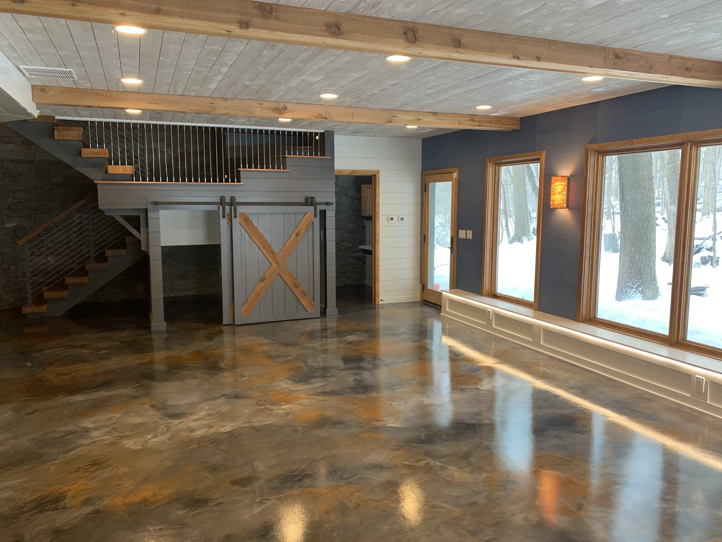 The glossy metallic epoxy flooring, shiplap ceiling with exposed wood beams, custom barn door beneath the floating staircase, and large viewing windows are bespoke features marking this walkout basement as part of a Booras Builders custom-built home.