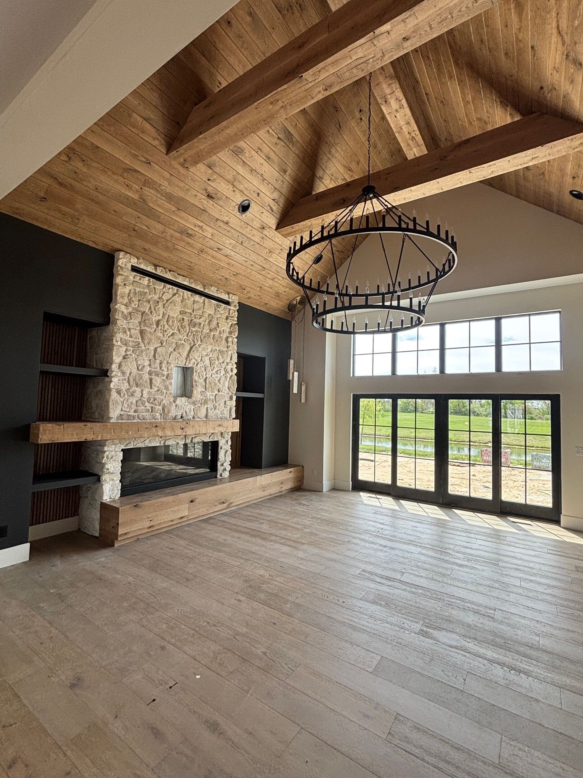 A traditional living room with a stone fireplace and a natural, wood-paneled raised ceiling with huge wood beams stretching across it. Tall windows provide natural lighting and an aesthetic view to this Porter County home built by Booras Builders.