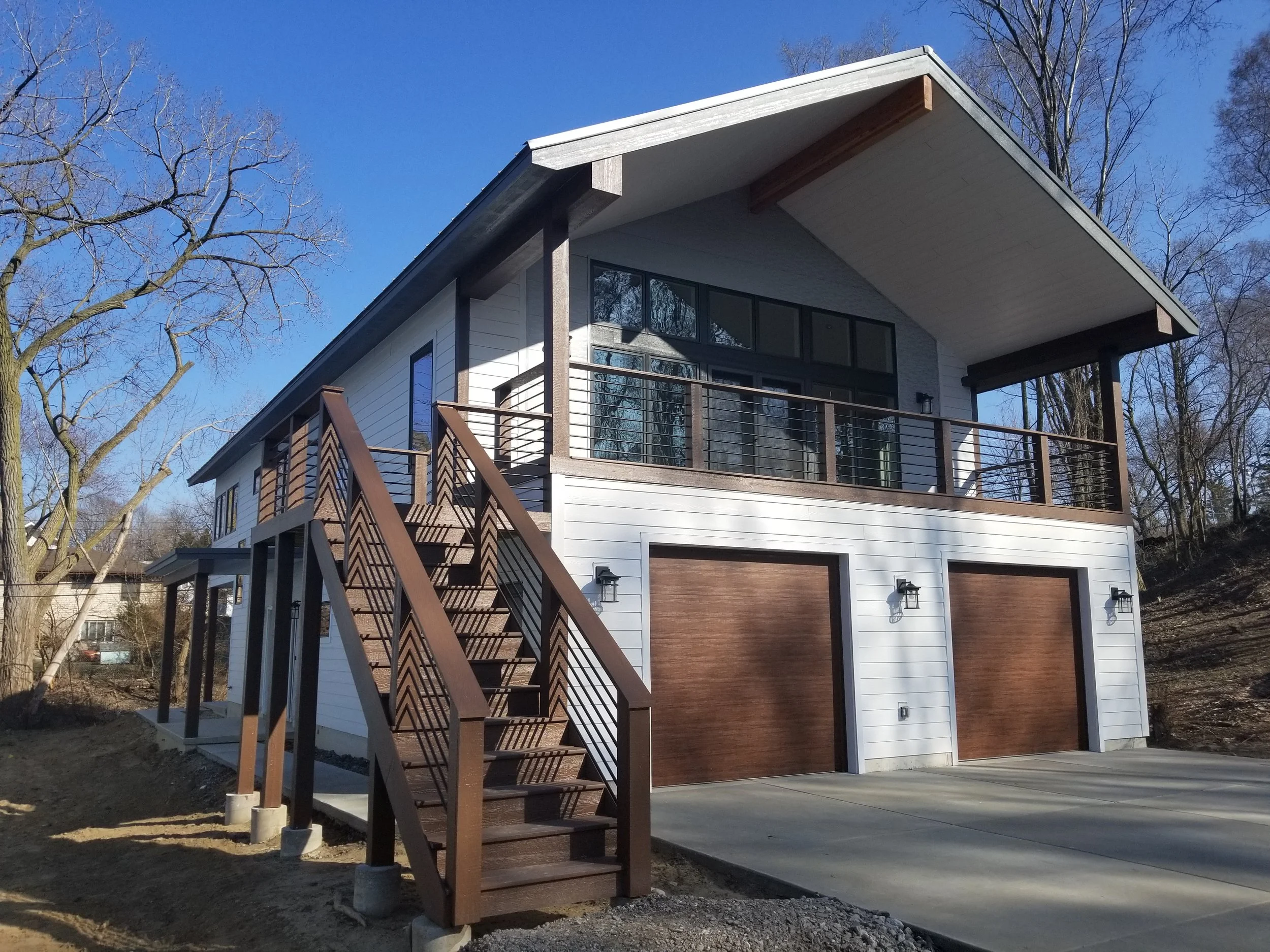 Designed around bold geometric angles, this contemporary custom home pairs a dramatic gabled roof with expansive glass, an elevated front terrace, cable railings, and wood-accented garage doors. It was constructed by Booras Builders in Chesterton.
