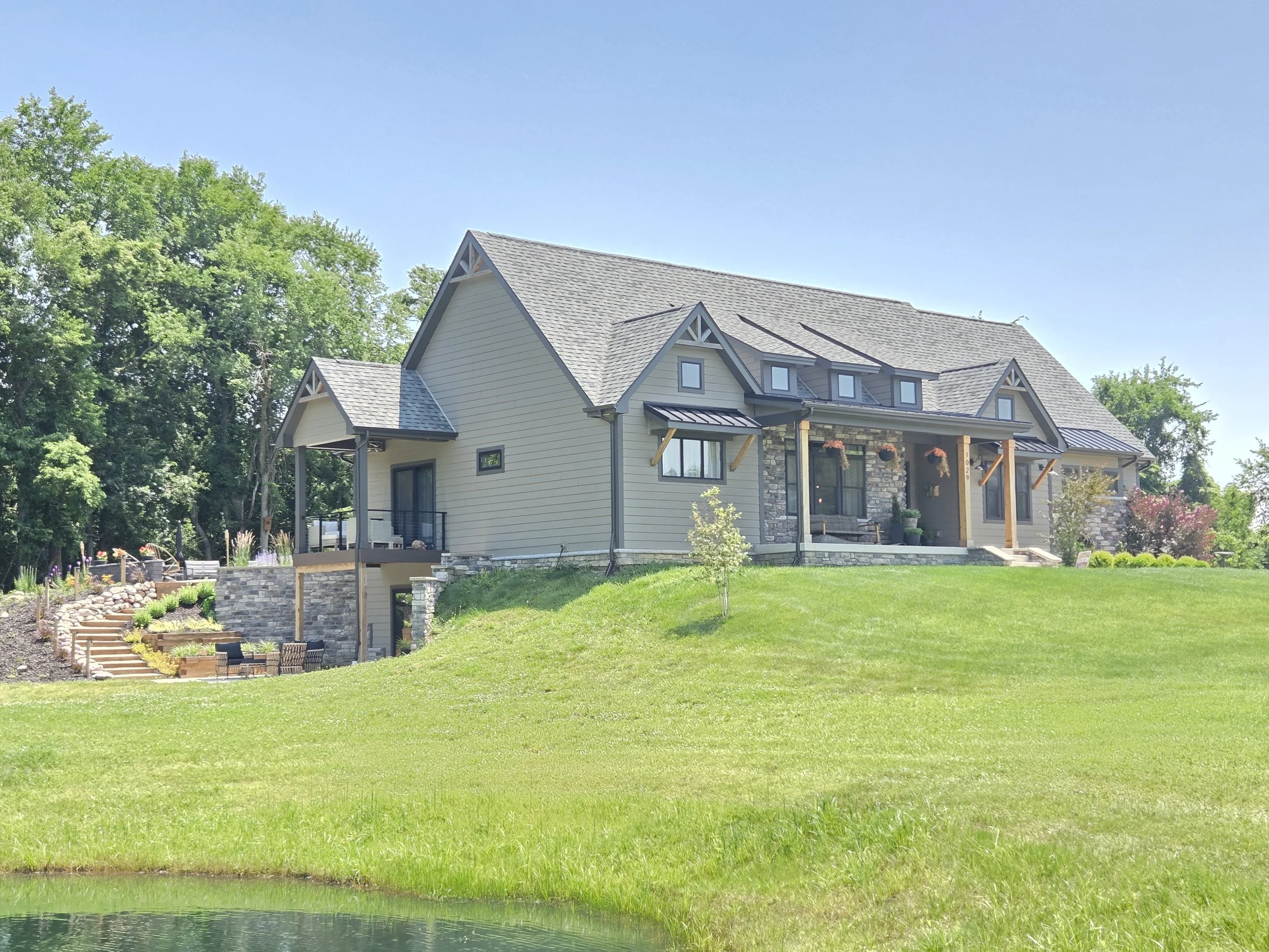Built on a sloped lot in Chesterton, this craftsman home features a walkout basement patio, stone veneer accents, three shed dormers, premium timber frame porch details, and landscaping overlooking a scenic pond view.