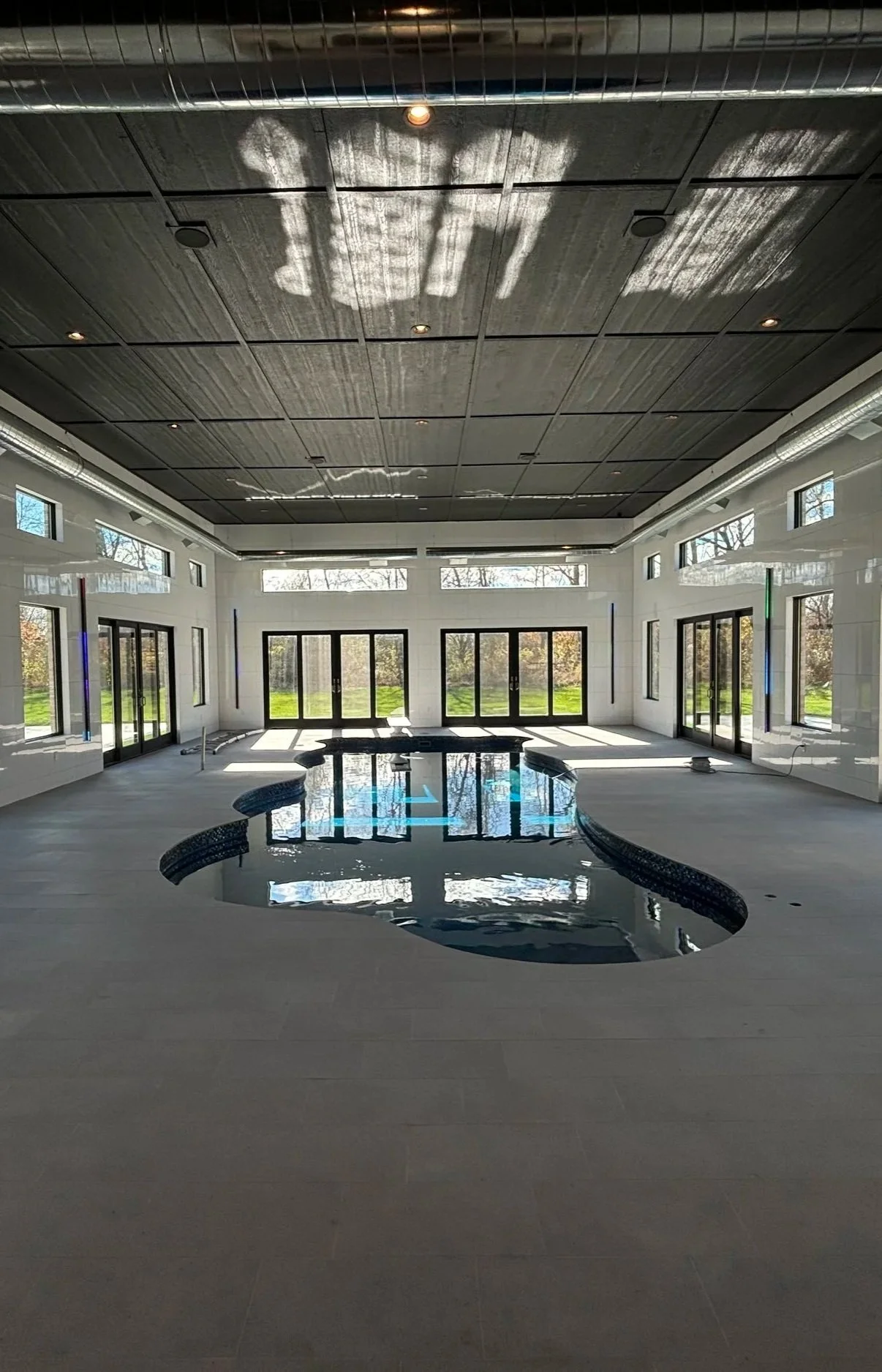 Surrounded by expansive glass doors and transom windows, a custom indoor pool with a dark finish anchors this sprawling pool house, showcasing tile flooring, white walls, and a dark paneled ceiling.