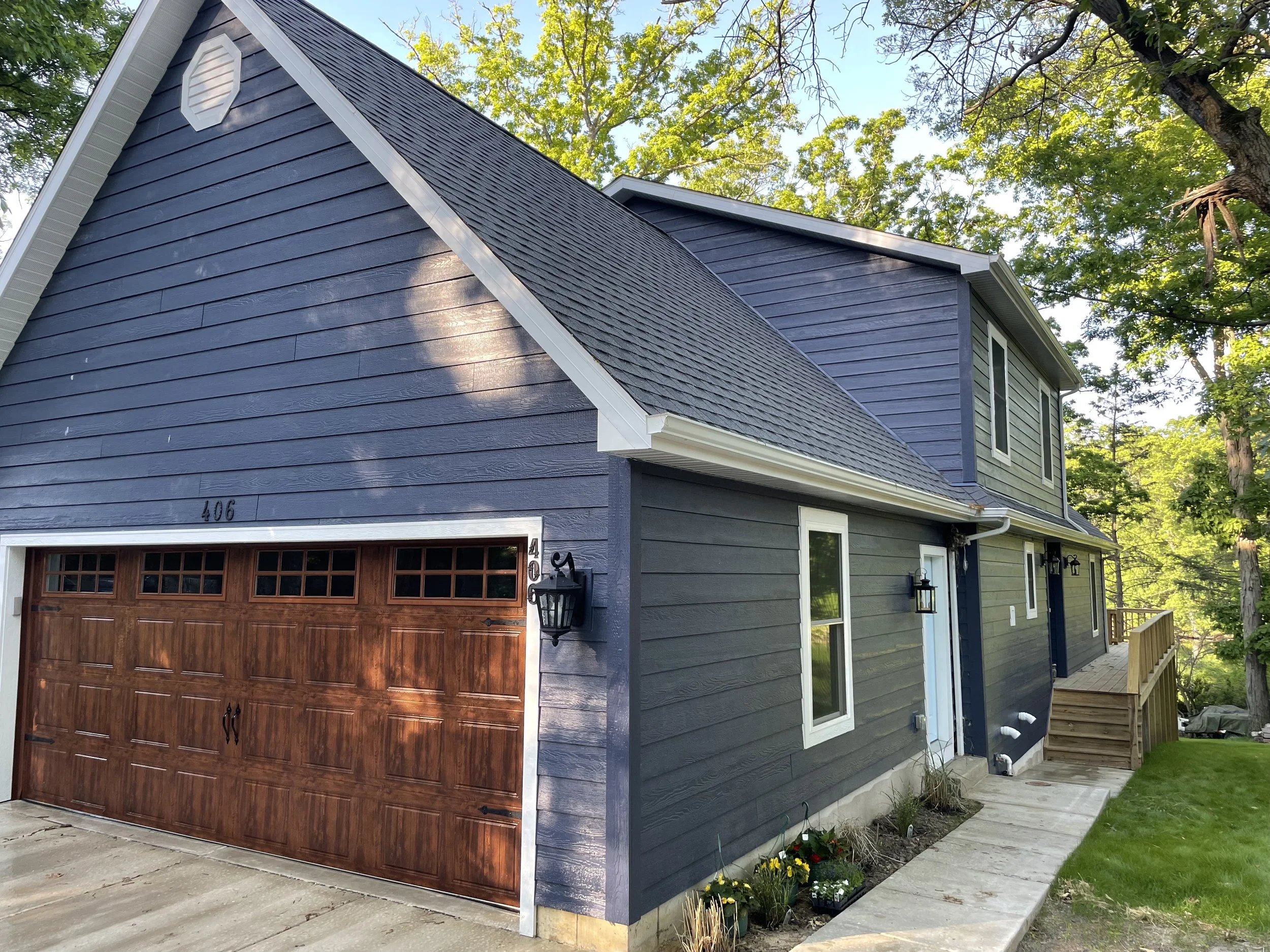 A Mid-Century home with gray-blue siding, a rustic-brown colored garage door, and fancy garage lights in Porter County. The Booras Builders crafted home is located in a wooded area.