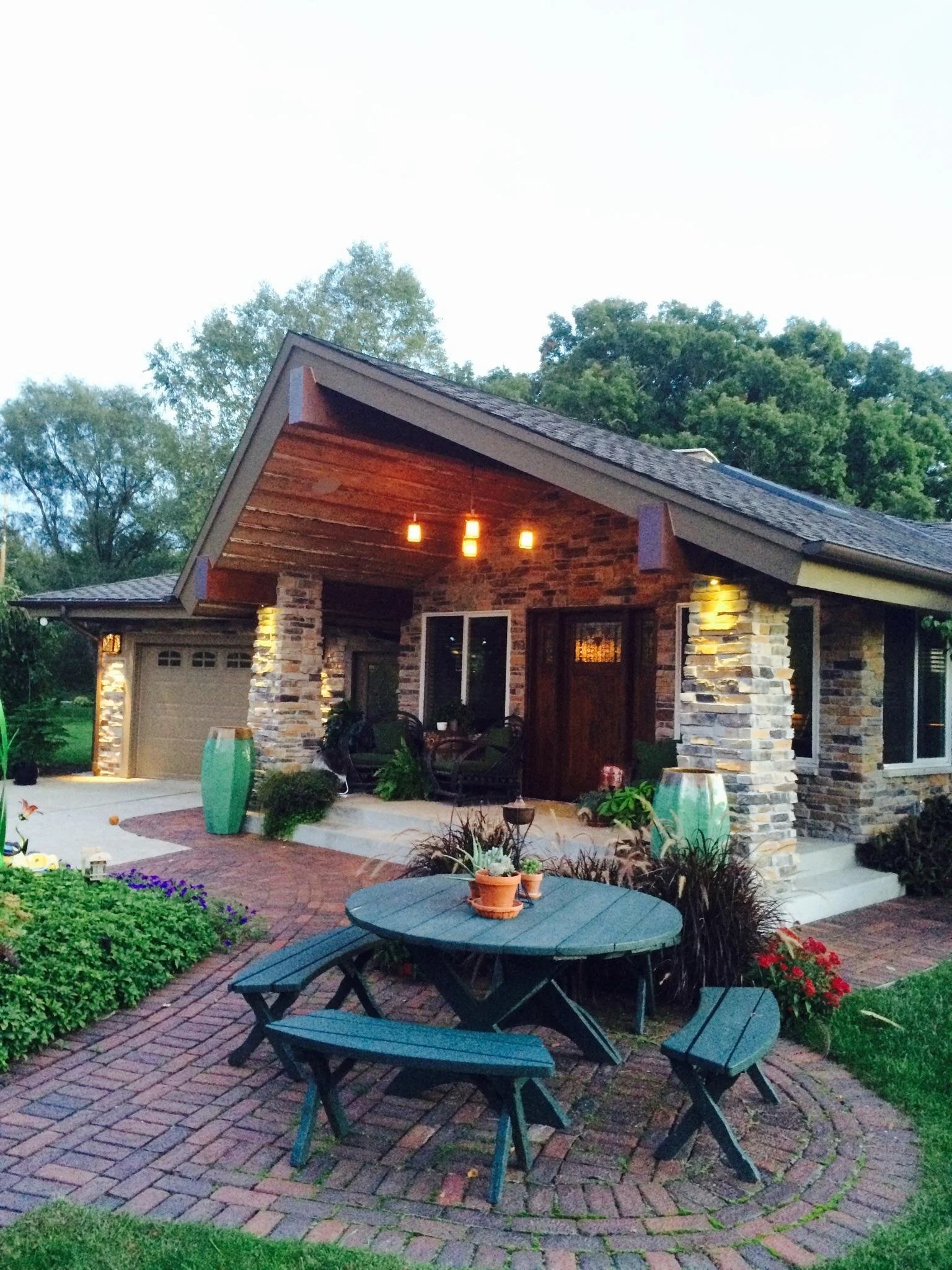 A vaulted porch roof, natural stone masonry, warm exterior lighting, a wooden front door, brick paver walkways, outdoor dining area, an attached garage, and pretty landscaping are all highlights of this inviting entrance to a custom-crafted residence
