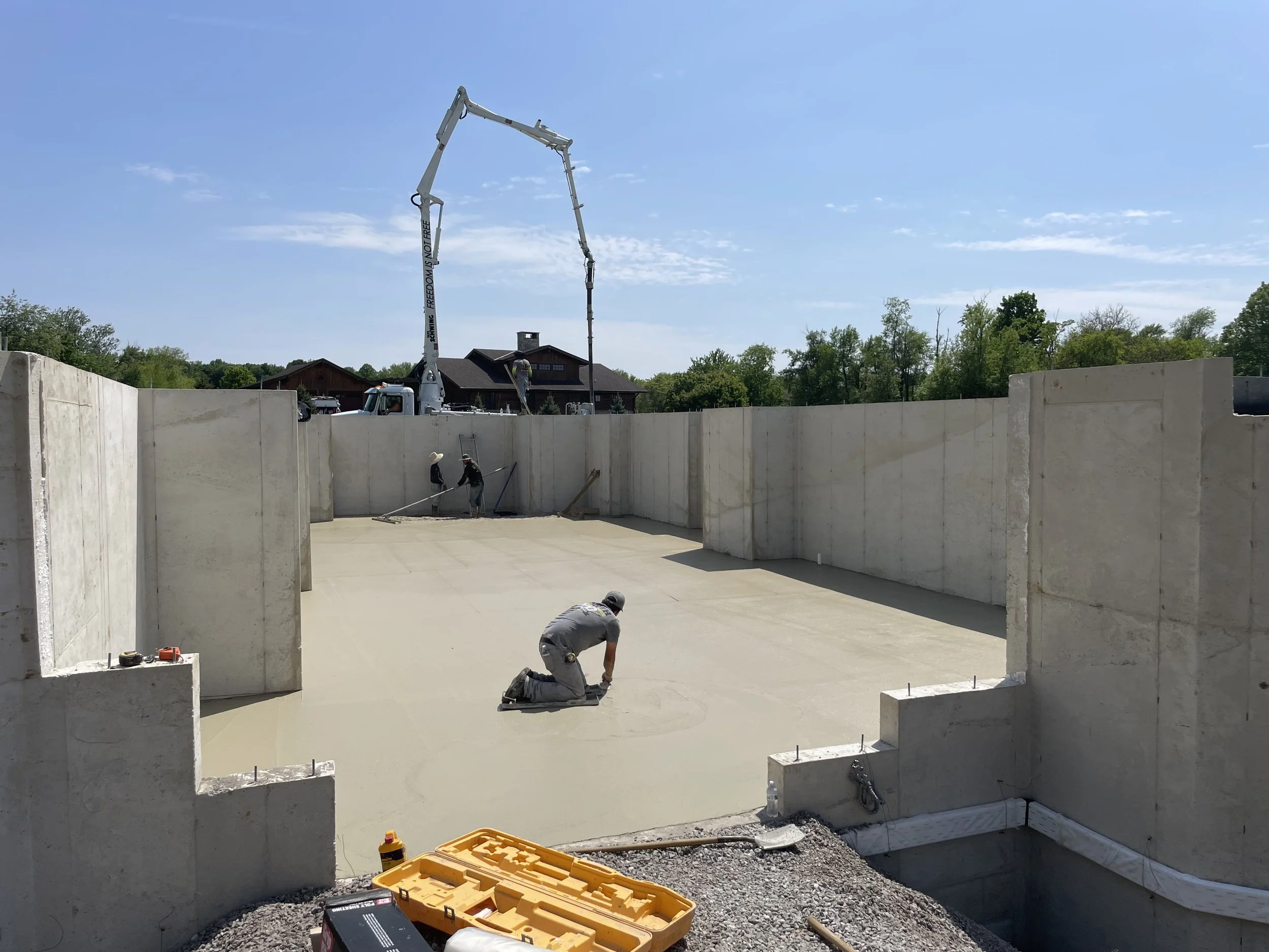A concrete crew completes the smooth trowel finish on a freshly poured basement floor slab of what will be a Booras Builders custom-built home, surrounded by tall poured concrete foundation walls beneath a boom pump truck operating on a clear day.