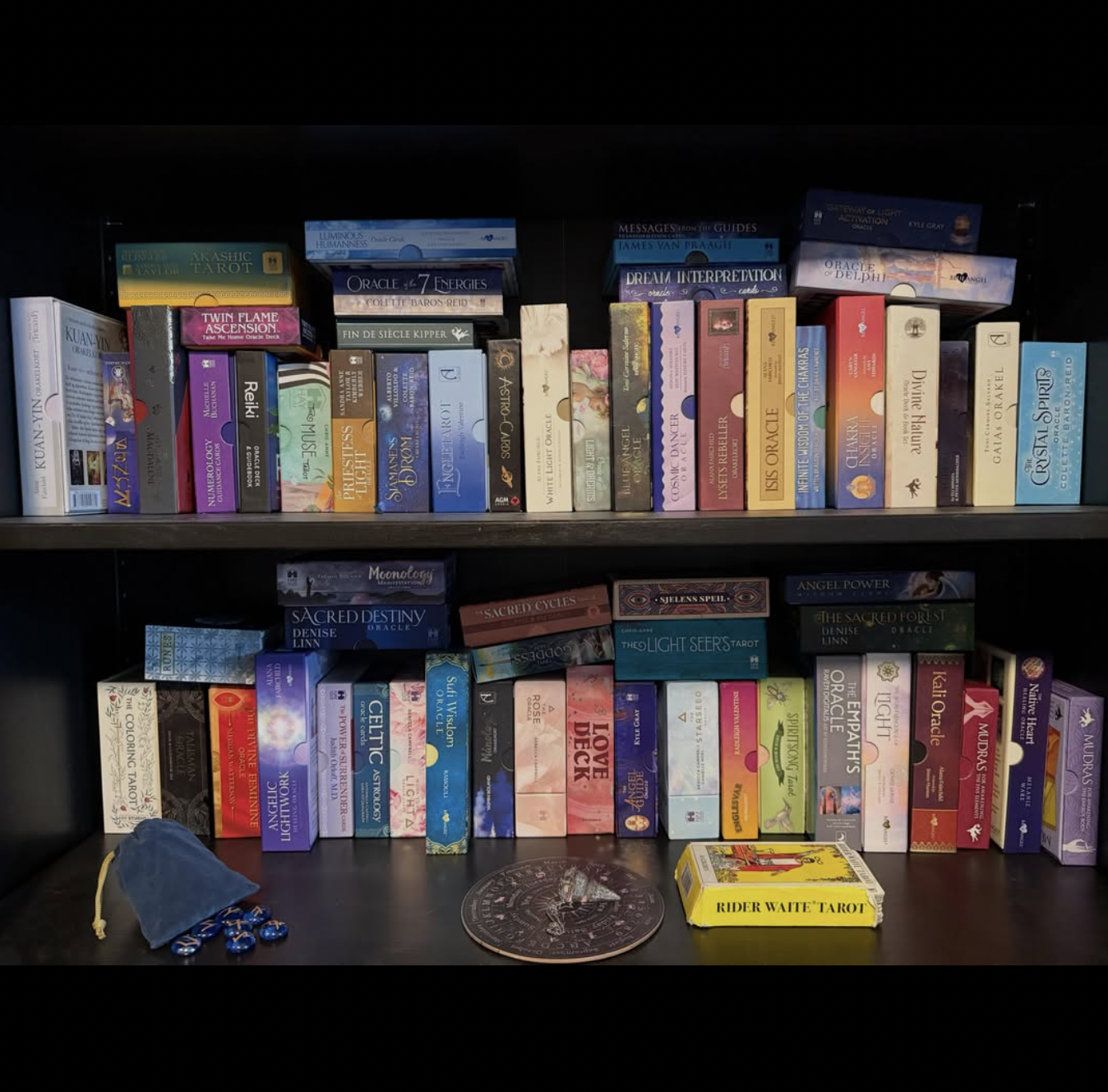 A bookshelf filled with tarot and oracle card decks, along with some accessories like small stones, a drawstring bag, and a Rider Waite tarot deck box, all organized on two shelves.