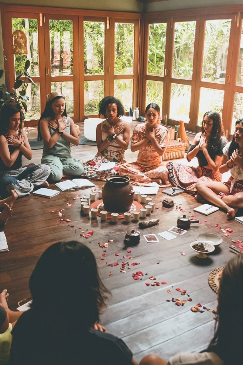 A group of women sitting cross-legged in a circle around a central pottery jar on a wooden floor, with some books, candles, and flower petals, in a sunlit room with large windows and greenery outside, engaged in a meditation or spiritual ceremony.
