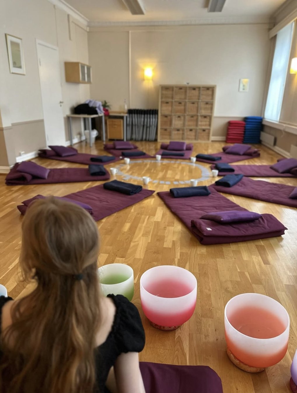 A young girl sitting on the floor of a meditation room with crystal singing bowls in front of her. The room has yoga mats and pillows arranged in a circle, with cups and blankets on each mat, and windows letting in natural light.