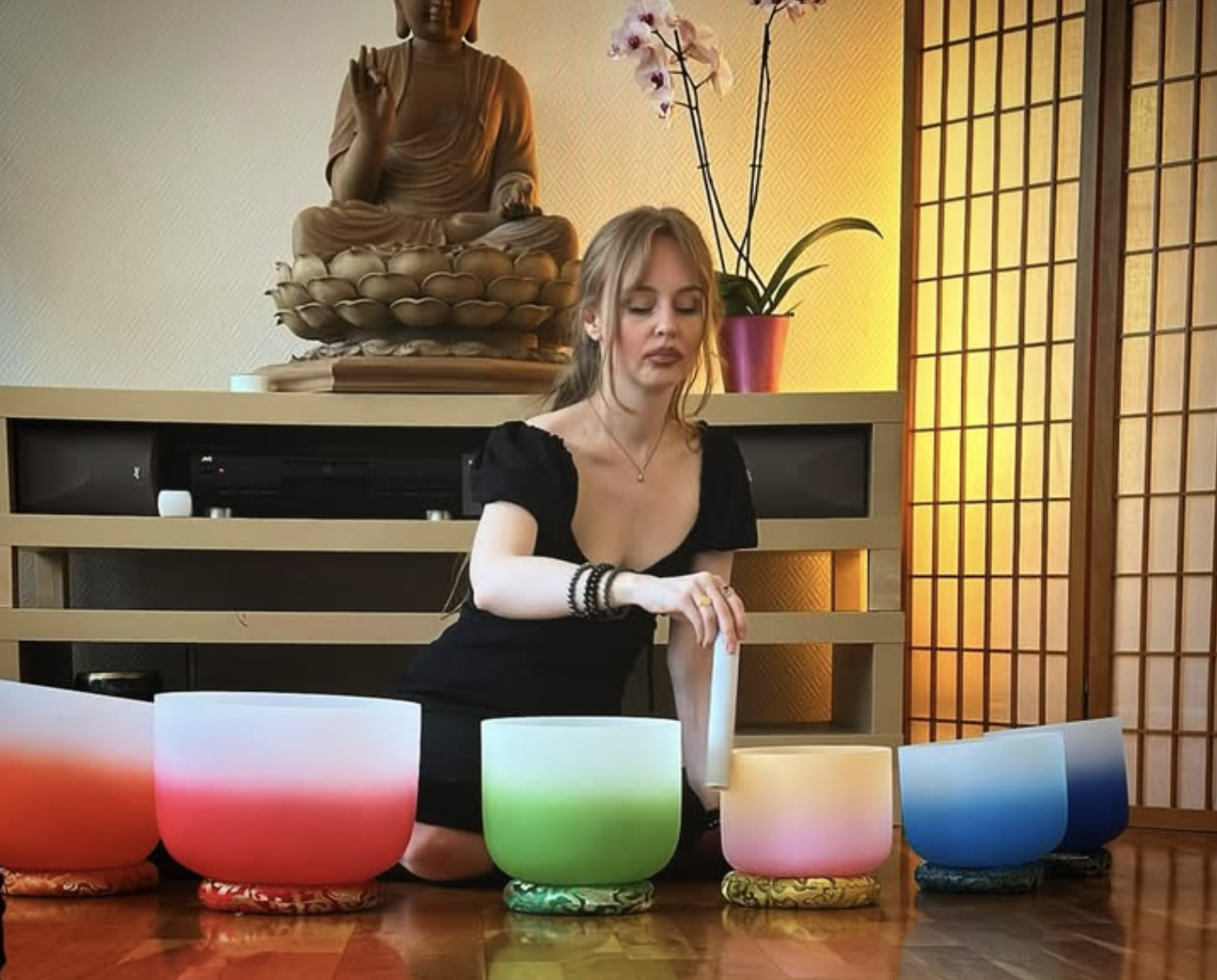 A woman dressed in black kneeling on a wooden floor, playing colorful singing bowls arranged in a row. Behind her is a wooden shelf with a Buddha statue, a potted orchid, and a decorative room divider.