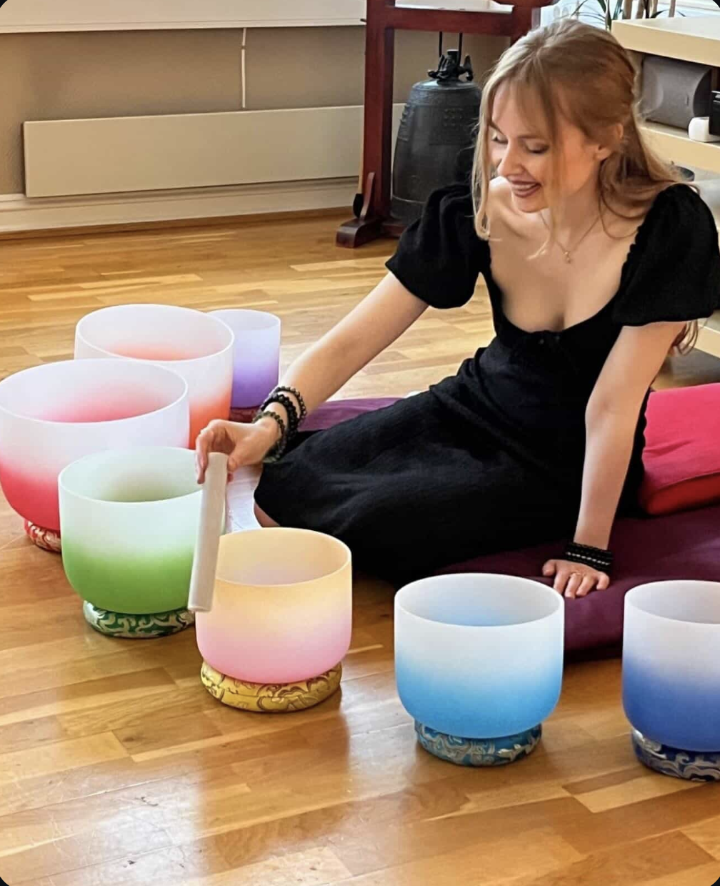 A woman with long red hair, wearing a black dress, sits on a purple mat and plays a set of colorful singing bowls with a mallet on a wooden floor.