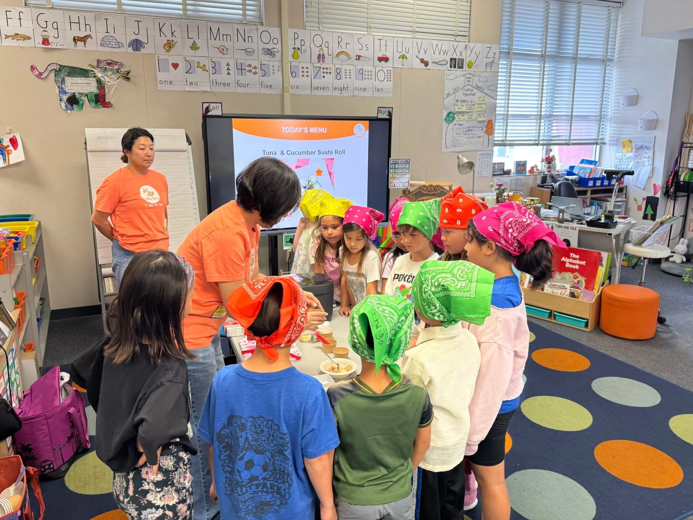 A classroom with children wearing colorful bandanas around their heads. Two women are supervising them. The children are gathered around a table, participating in a food activity with bowls of food. There is an electronic screen displaying a menu, and the classroom walls are decorated with alphabet charts and student artwork.