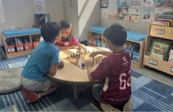 Three children playing chess at a small round table in a classroom, with educational posters and books on shelves in the background.