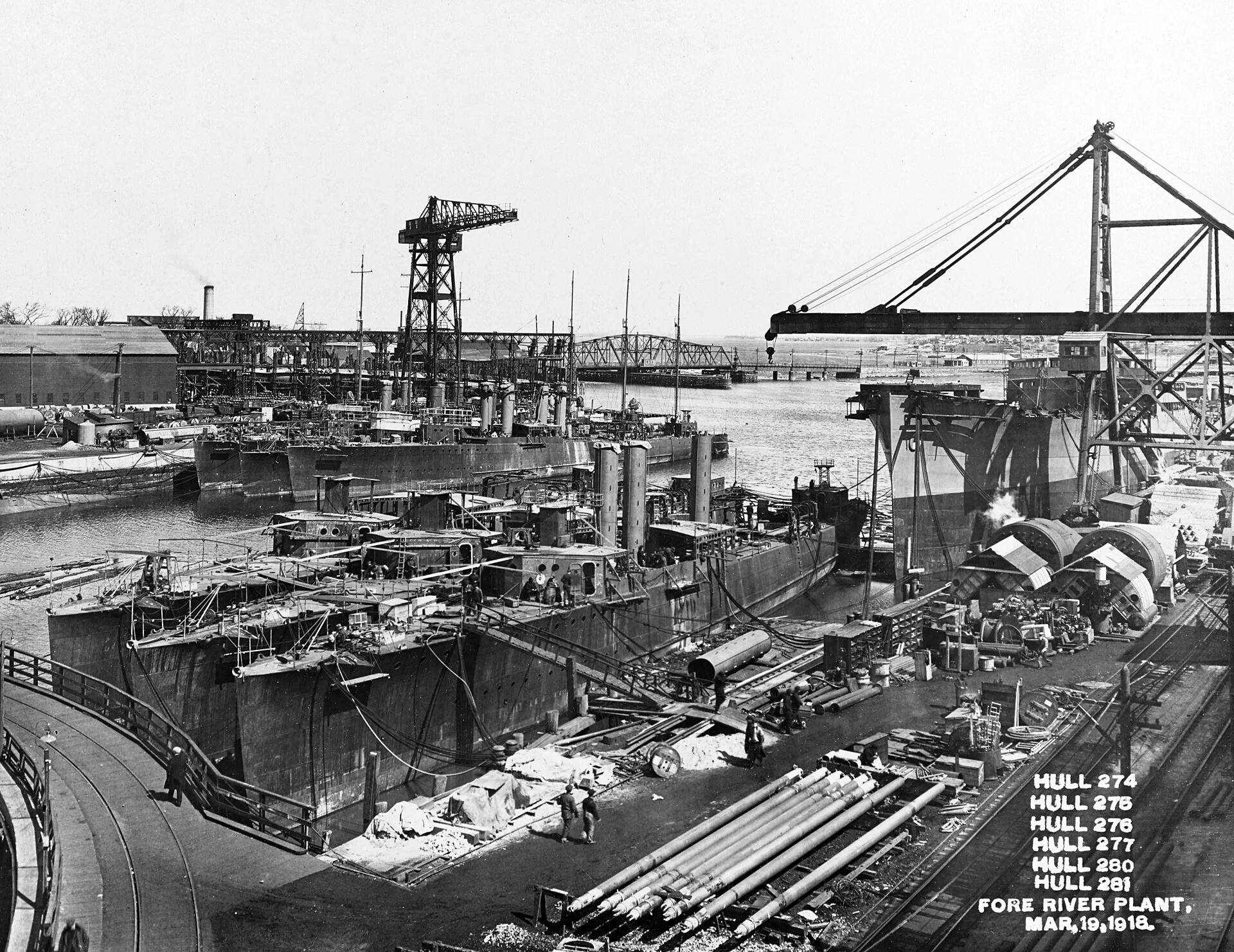 Black and white photo of a shipyard with large industrial ships and cranes, cityscape in the background, and a road running along the water's edge. Visible text in the bottom right corner indicates it's the Hull 274, a fore river plant, dated March 19, 1918.