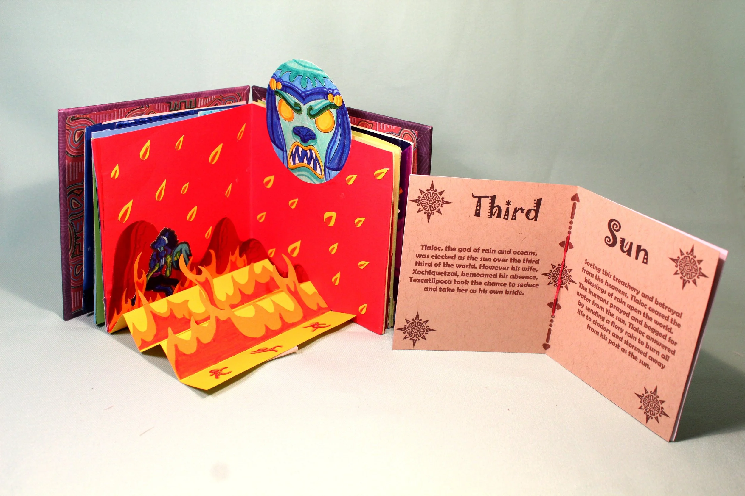 Colorful folded paper booklet with a red fire illustration, a painted mask with an angry expression, and text about the god Tlaloc on a pink page.
