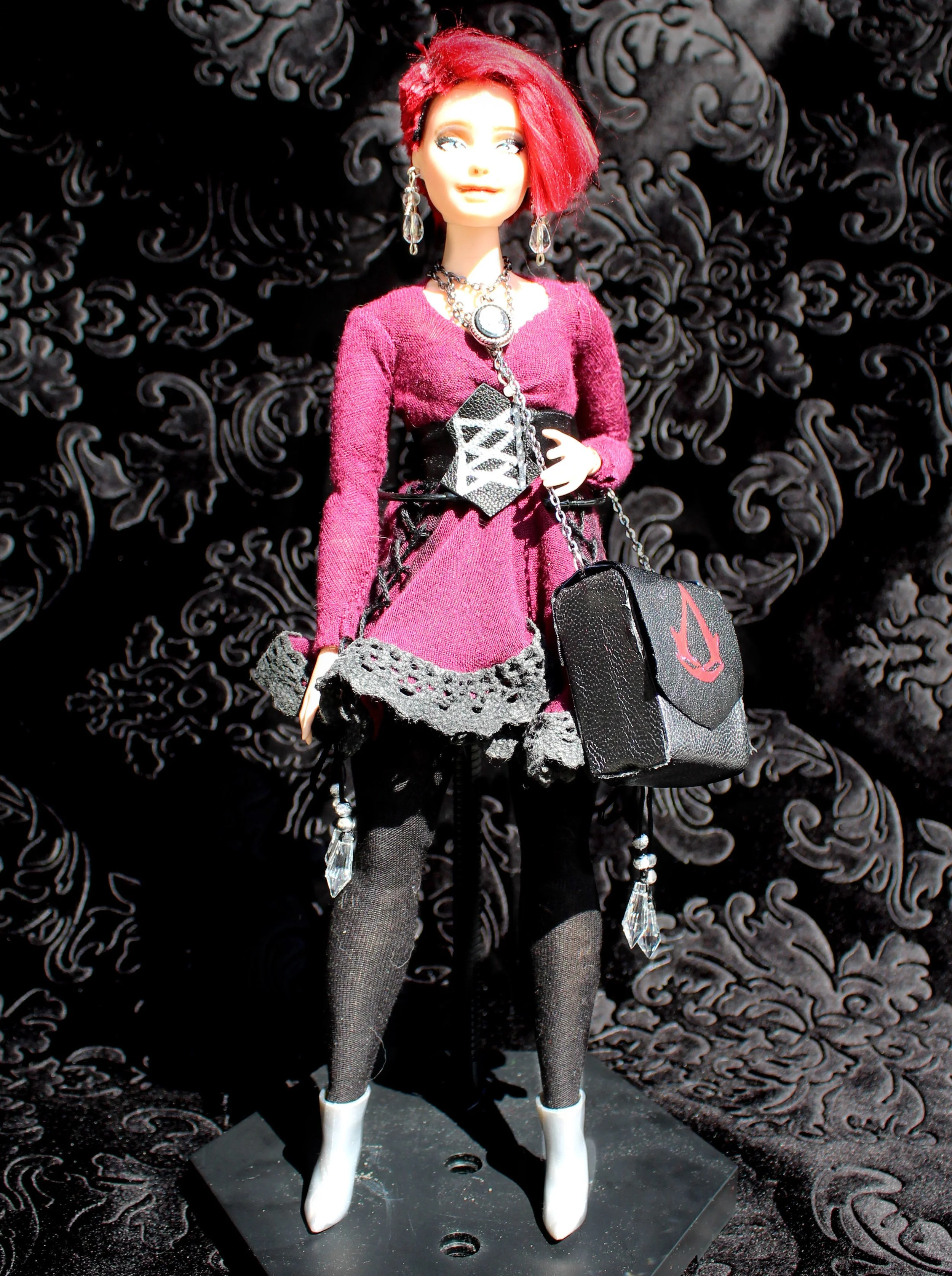 A fashion doll with short red hair, wearing a burgundy knit top, black lace leggings, and white high-heeled boots. She has jewelry including dangling earrings and layered necklaces, and is holding a black purse with a chain strap. The background feat