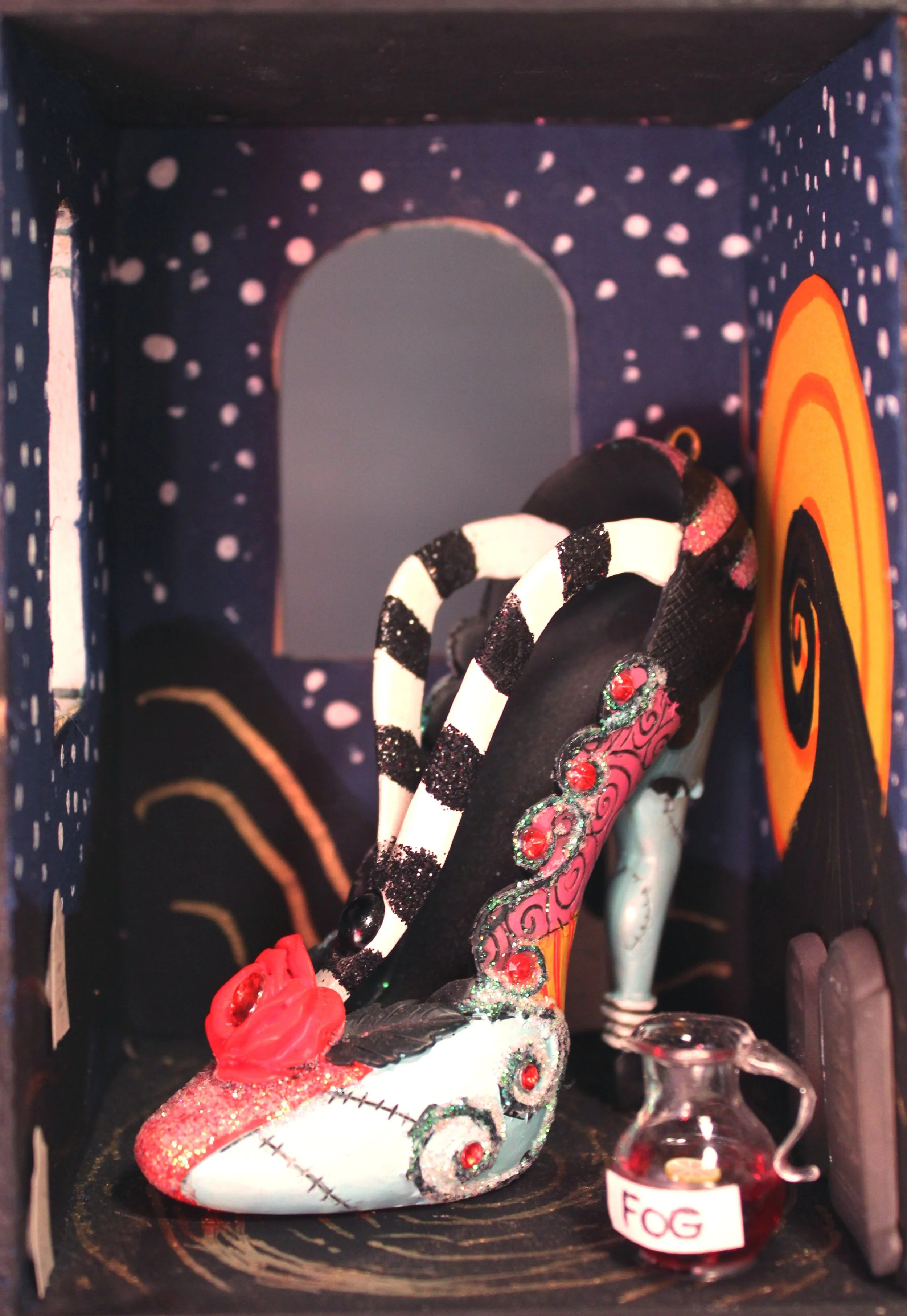 A decorative skull-shaped object with colorful and glittery designs, including red and black accents, positioned inside a box with a dark, starry background. A potion bottle labeled 'Fog' is in front of the skull.