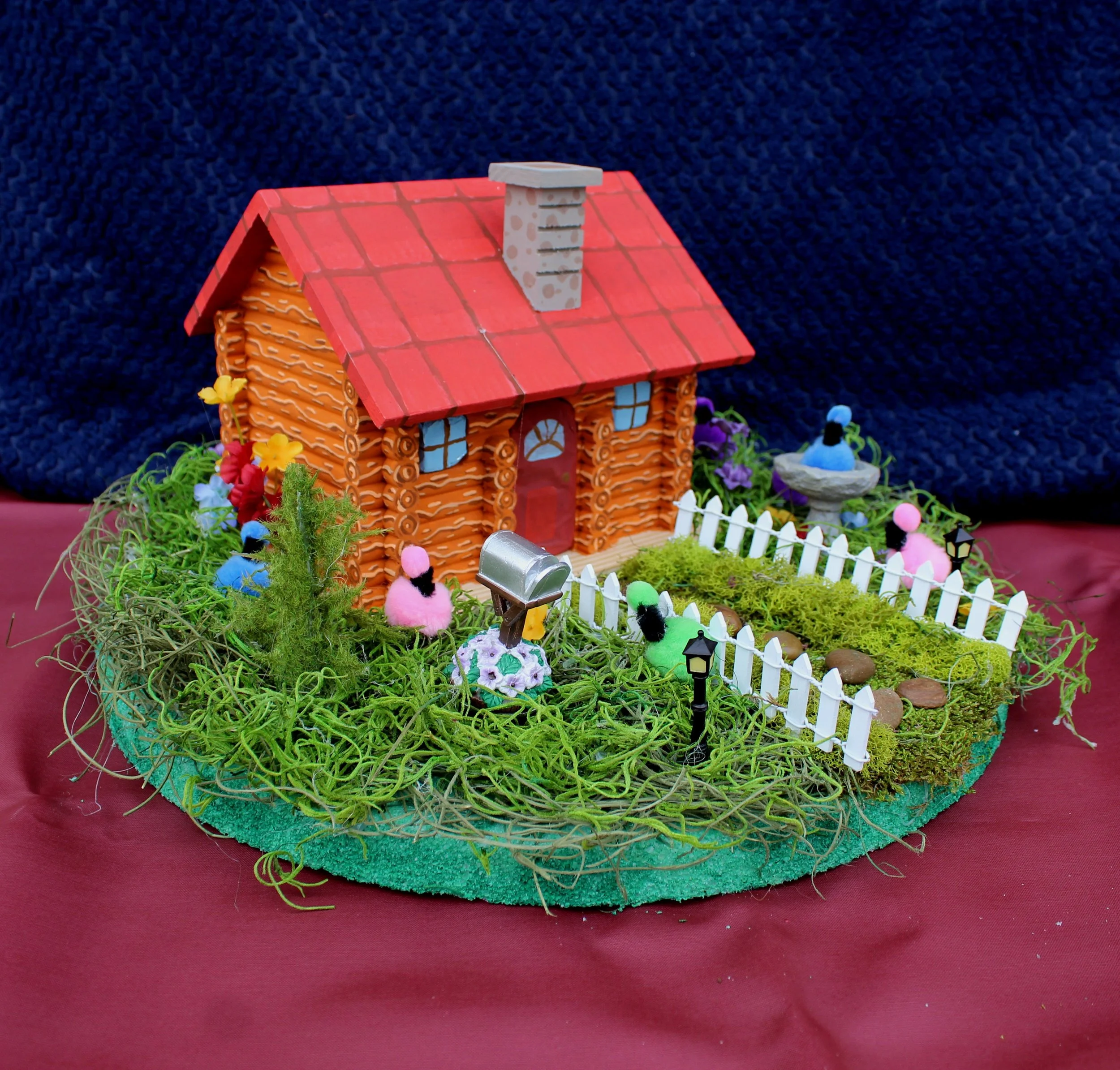 Miniature house with a red roof, blue window panes, surrounded by colorful Easter eggs, flowers, and a white picket fence on a green grassy base.