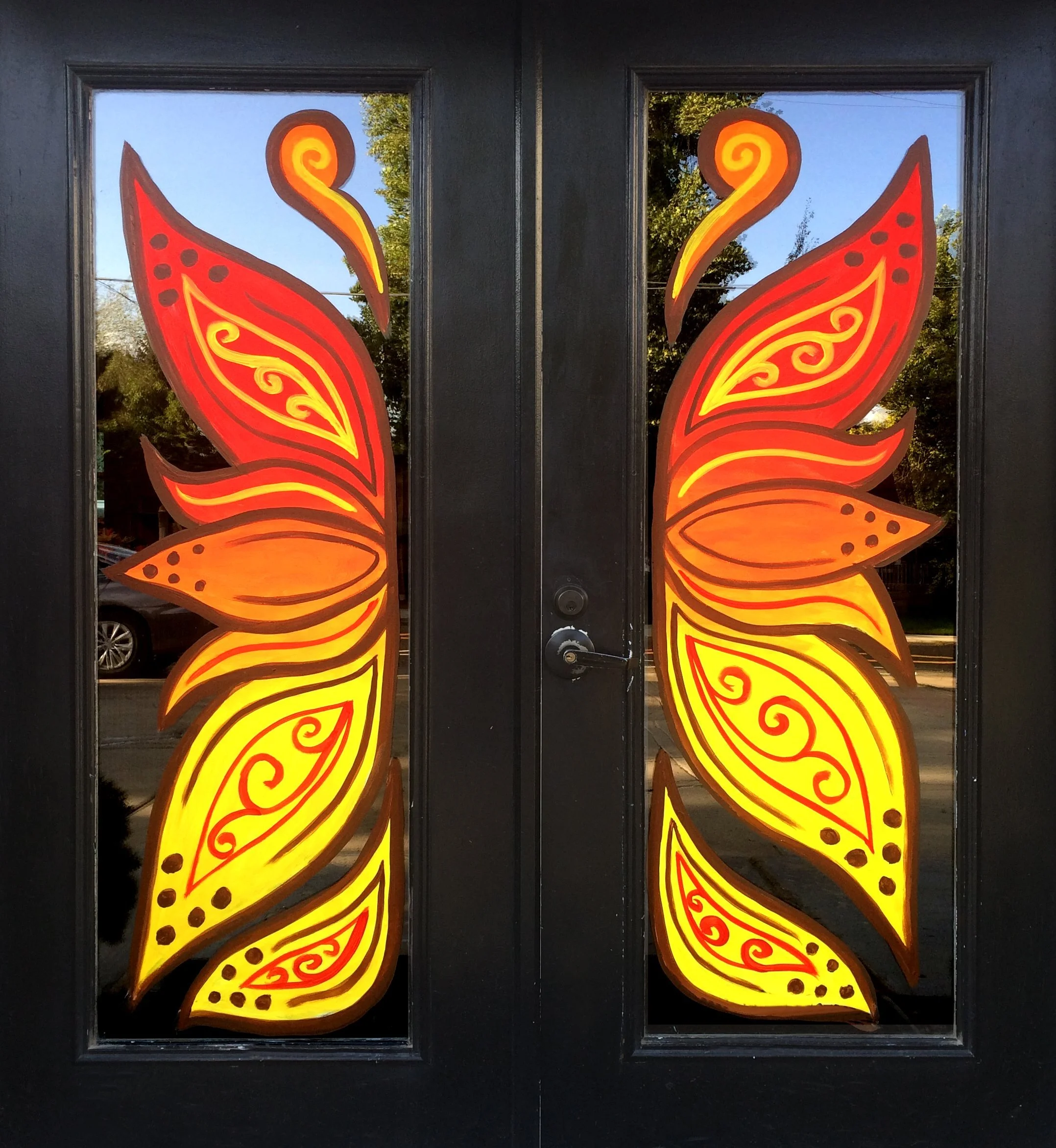 Decorative black door with illuminated butterfly wings painted on glass panels, with yellow, orange, and red swirl patterns, reflecting trees and blue sky outside.