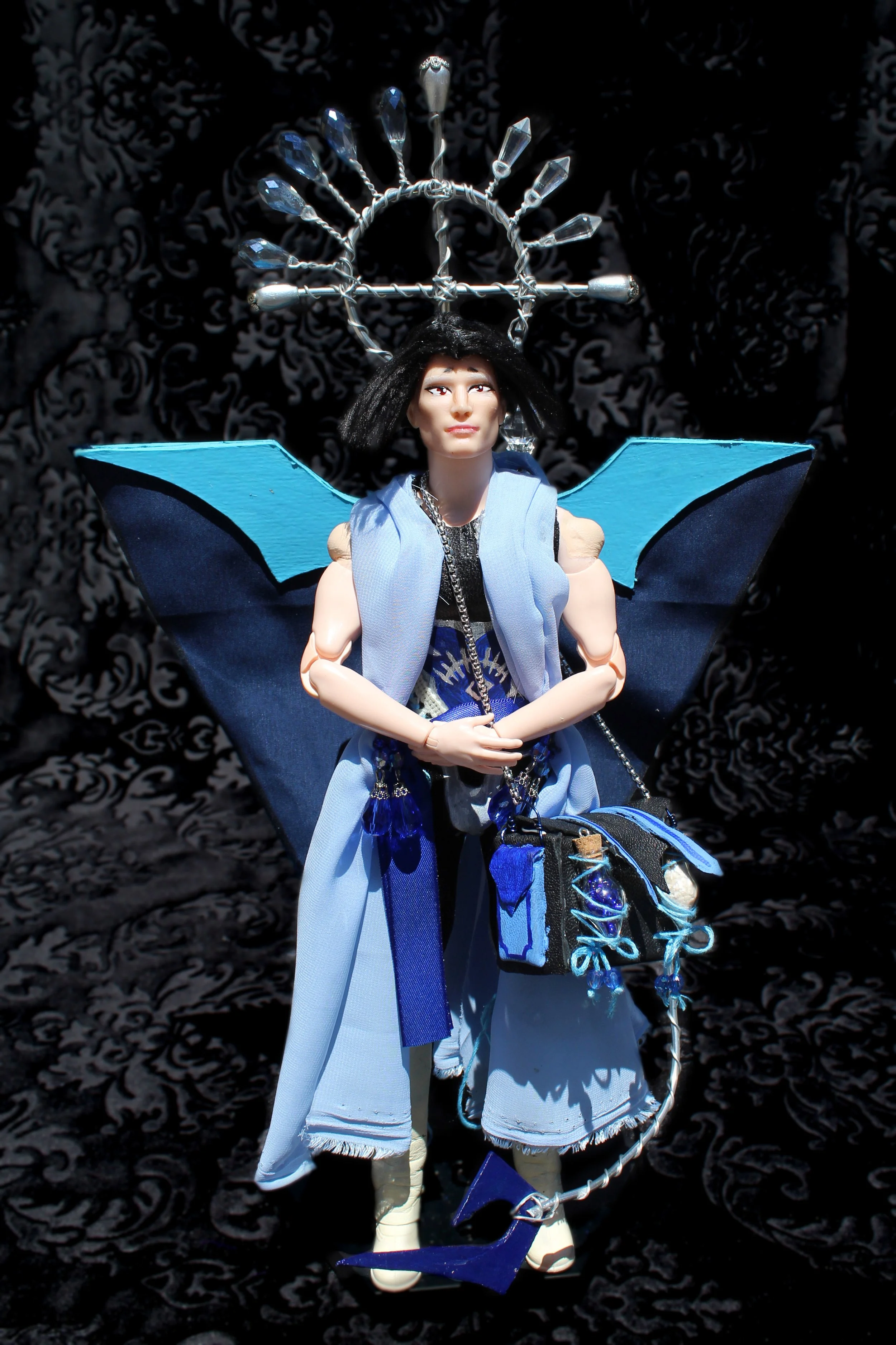 A doll with blue butterfly wings, wearing a blue and white outfit, has a headpiece with a metal structure and blue accents, set against a dark patterned background.