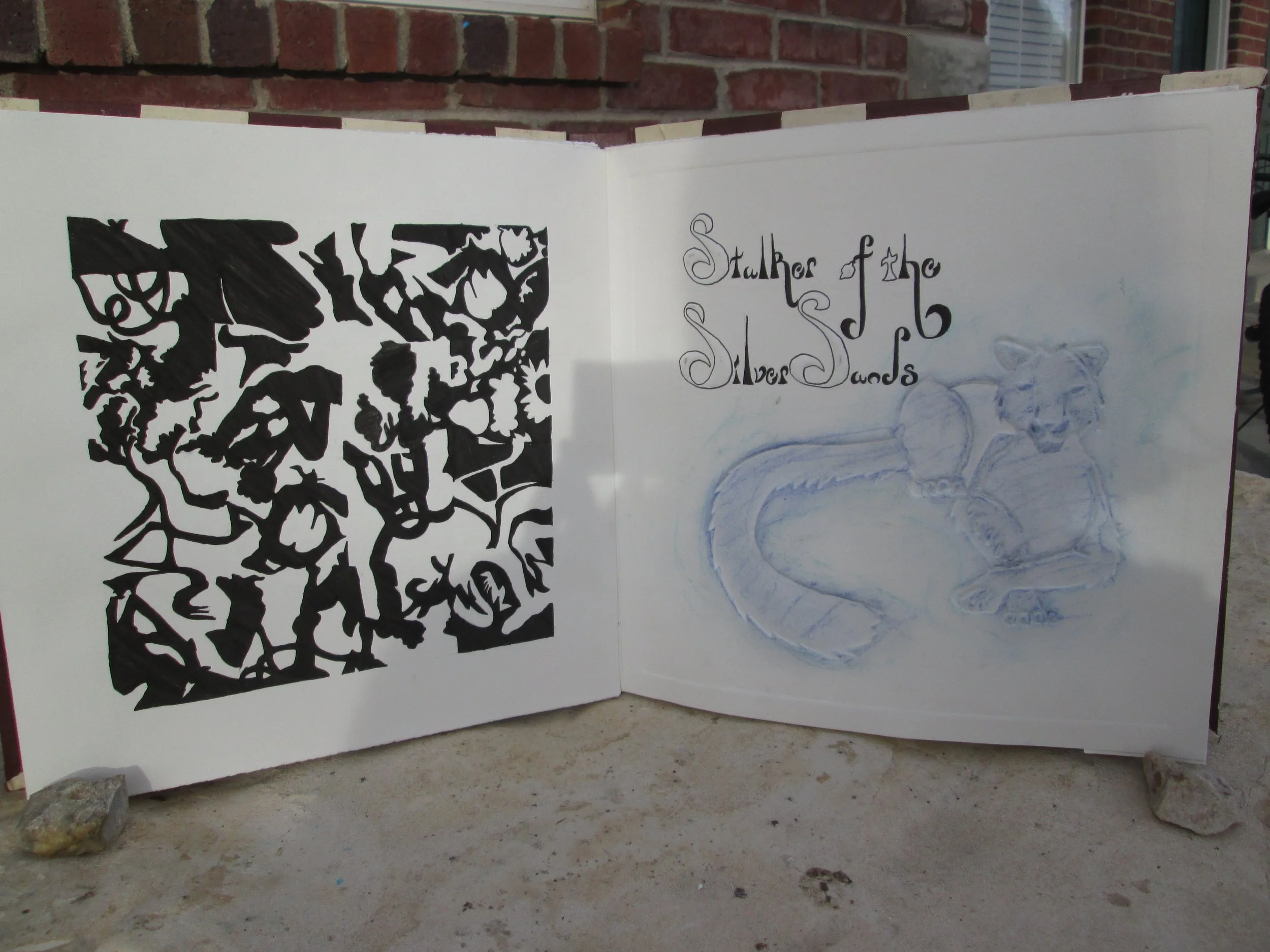 An open sketchbook with black and blue drawings and text, resting on a concrete surface with a brick building in the background.