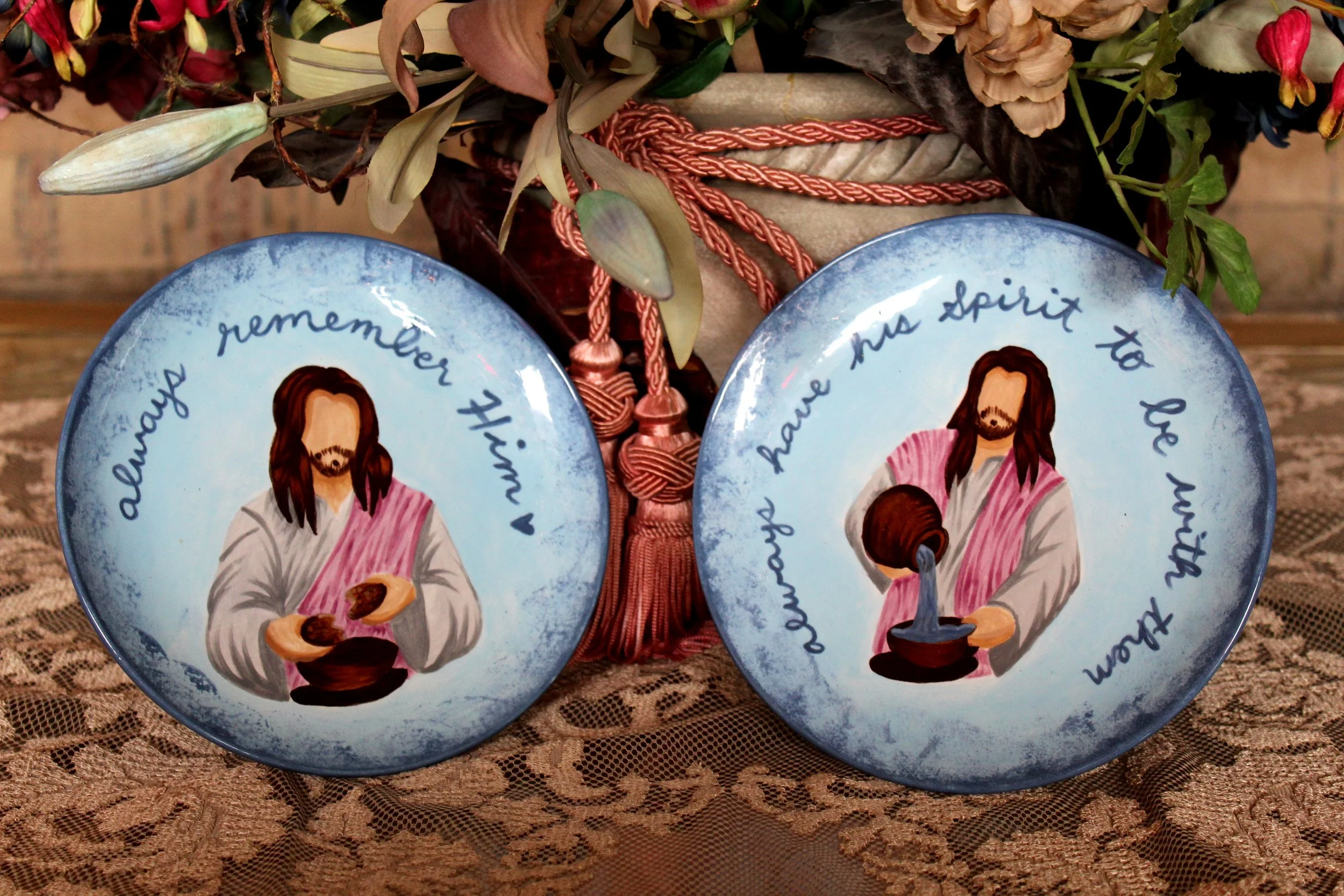 Two decorative plates with religious artwork of Jesus and a girl, with handwritten text around the images, placed on a lace tablecloth with a flower arrangement in the background.