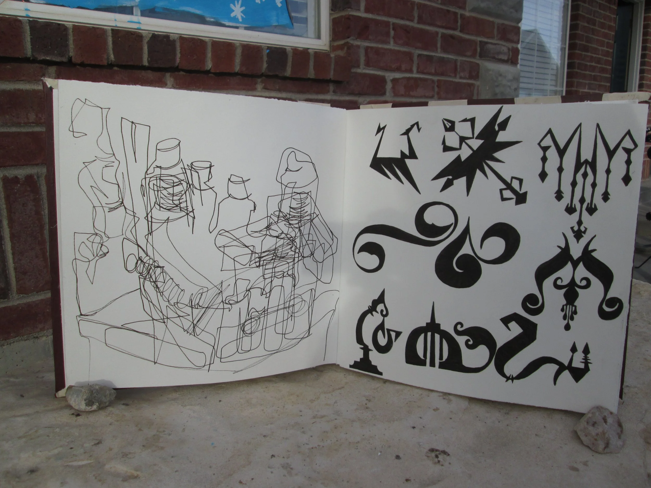 An open book with black abstract and decorative patterns on white pages is displayed outdoors against a brick wall. The left page features a chaotic line drawing, while the right page contains bold black designs.