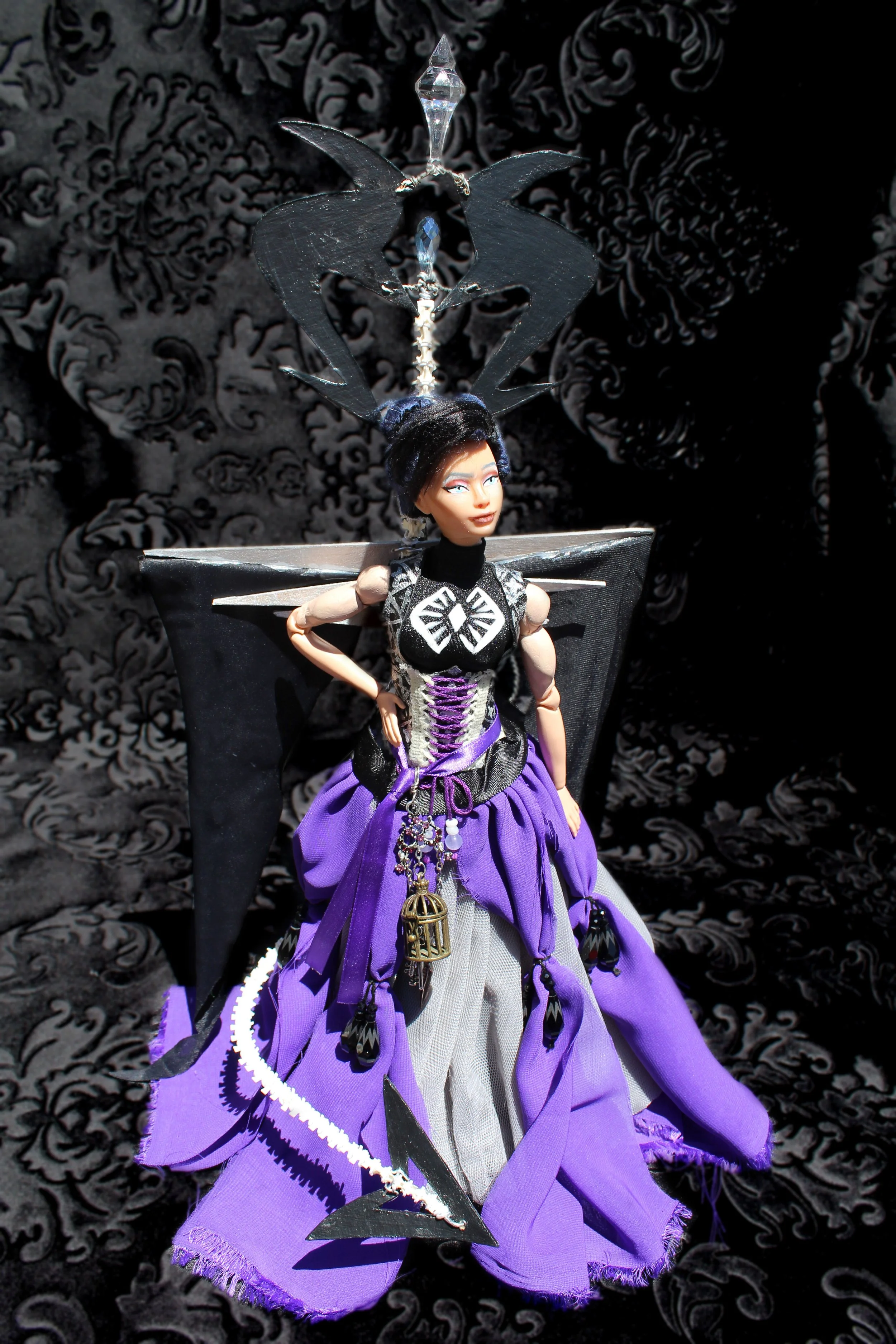A fashion doll wearing a purple and black dress with black wings, a headpiece with black and purple accessories, and a decorative staff, posed against a black patterned background.