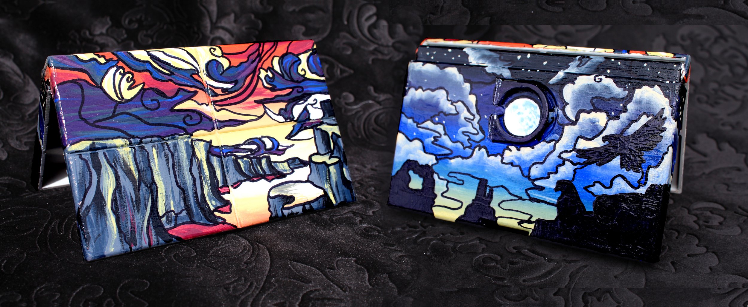 Two small, hand-painted, foldable canvases featuring colorful, abstract scenes with night sky themes, one with a sunset and the other with a full moon, placed on a black textured surface.