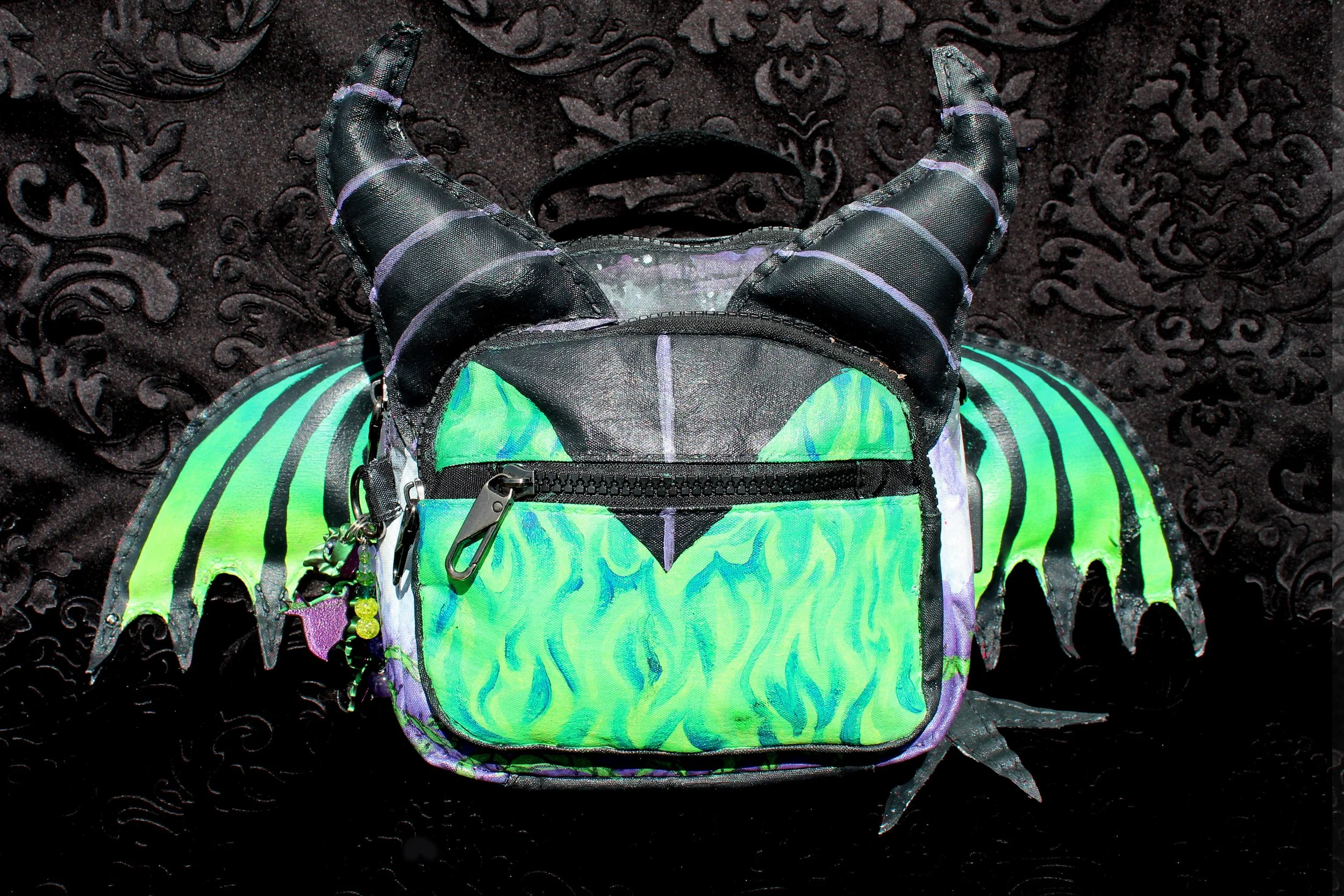 A black backpack with vibrant green and blue flame-like design, placed on a dark textured fabric background.