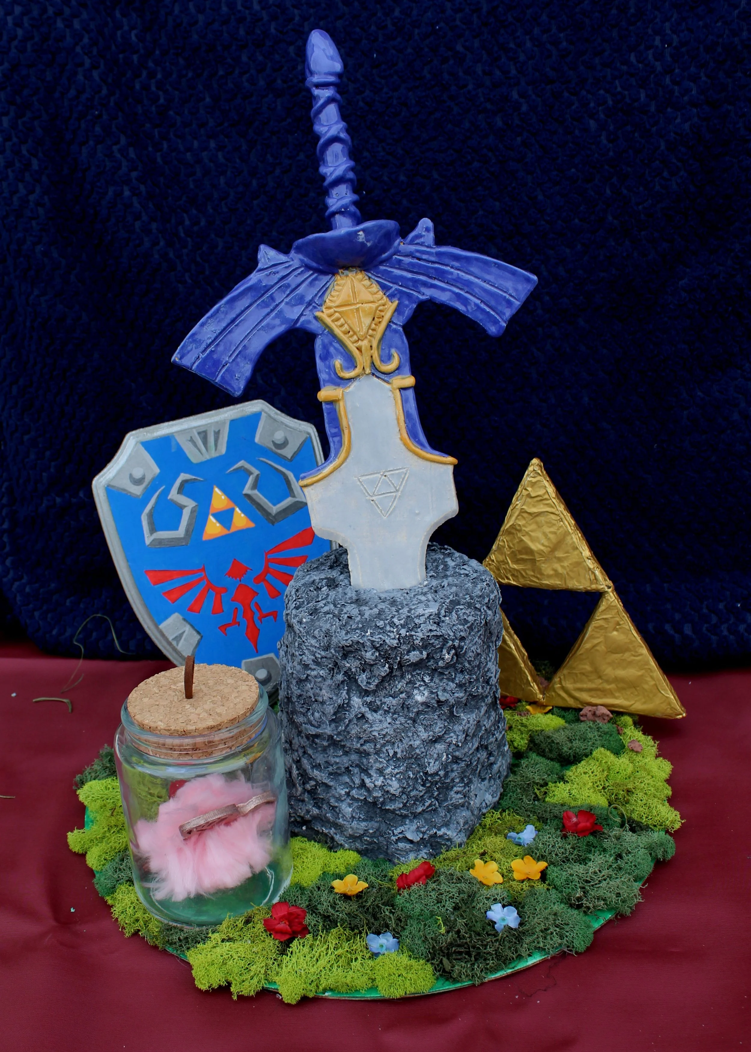 Decorative display featuring a stone-like base with a sword, a shield, and a golden cone, surrounded by colorful moss, small flowers, and various shapes on a red cloth background.