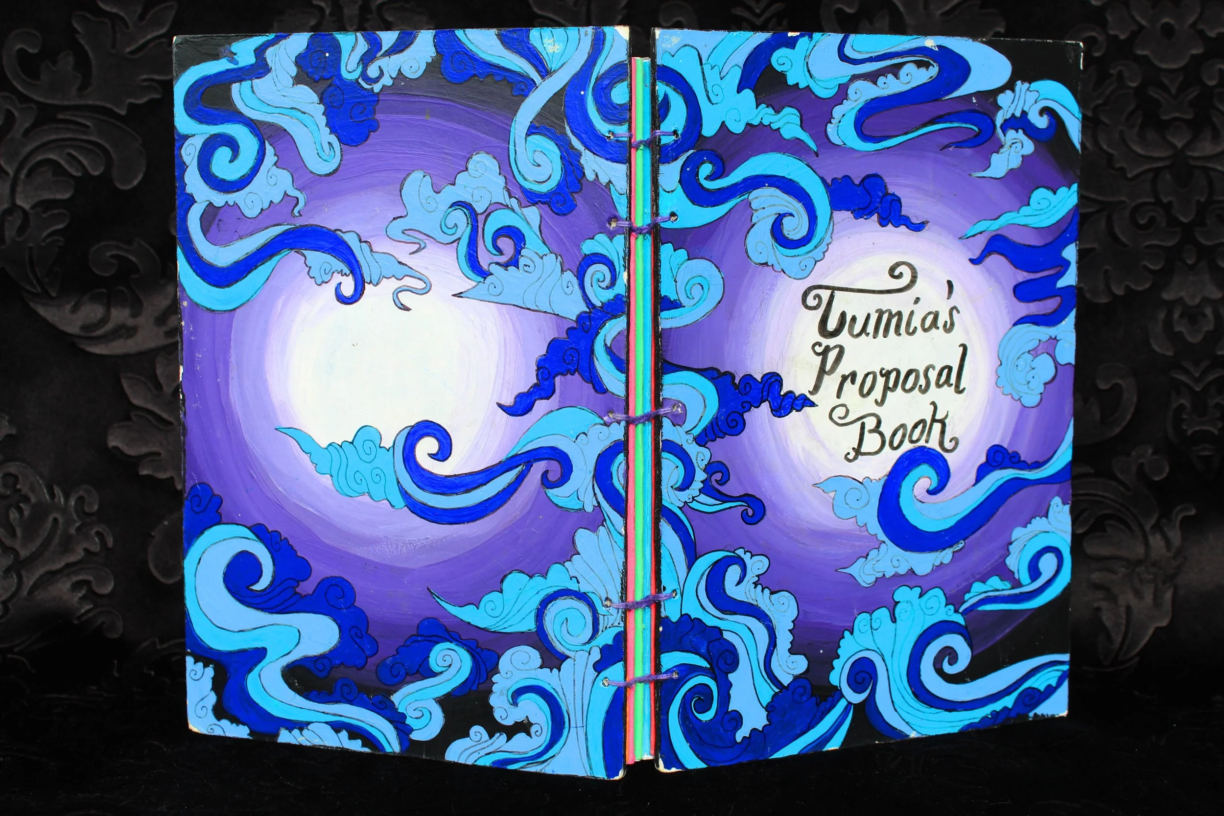 Colorful art or design featuring swirling blue clouds on a purple background with a central rainbow stripe and the text "Tumiás Proposal Book" written in black cursive.