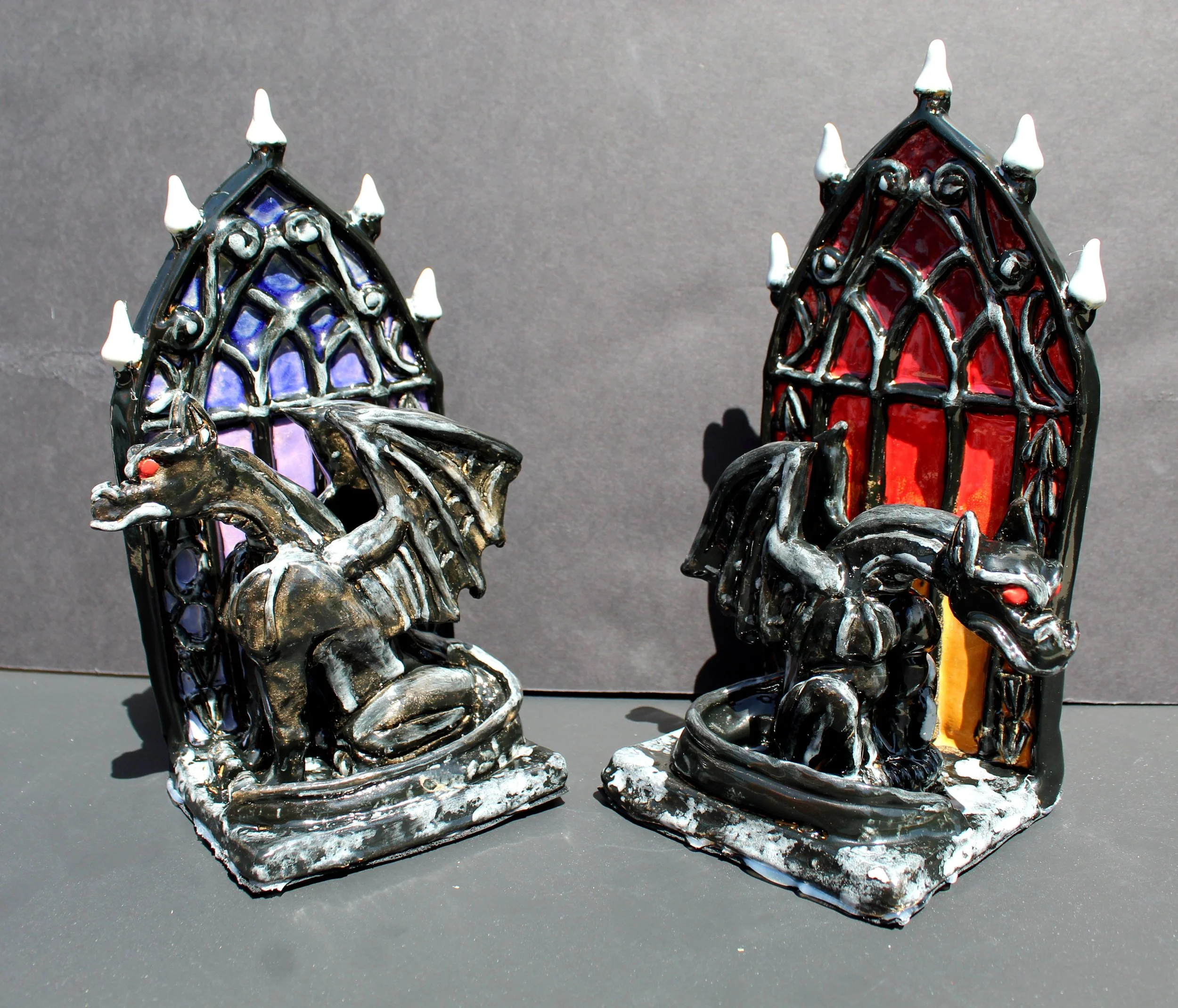 Decorative gothic-style metal candle holders resembling church windows with colored glass and small white candles on top, set against a gray background.