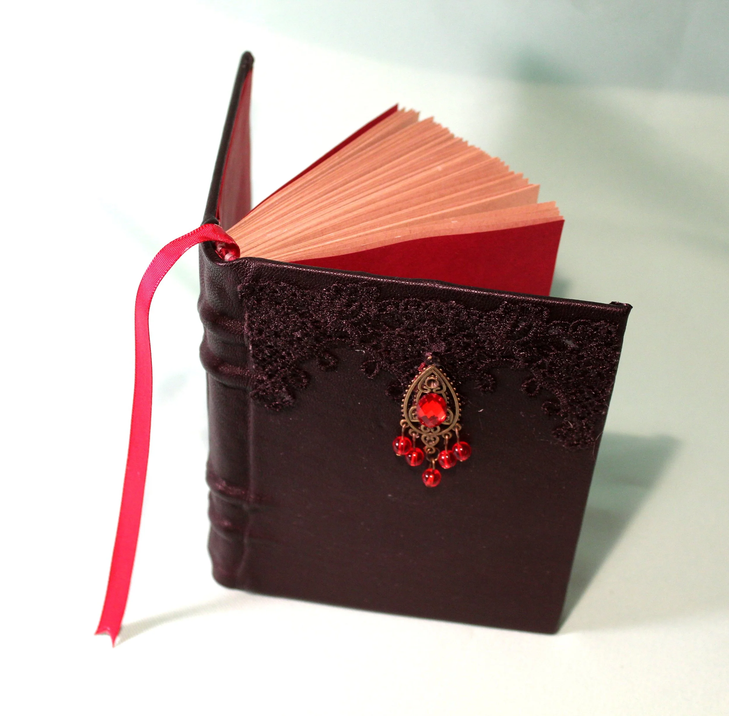 A burgundy leather-bound journal with decorative embroidery and a gold emblem with red accents on the front cover, open to reveal red pages and a pink ribbon bookmark.