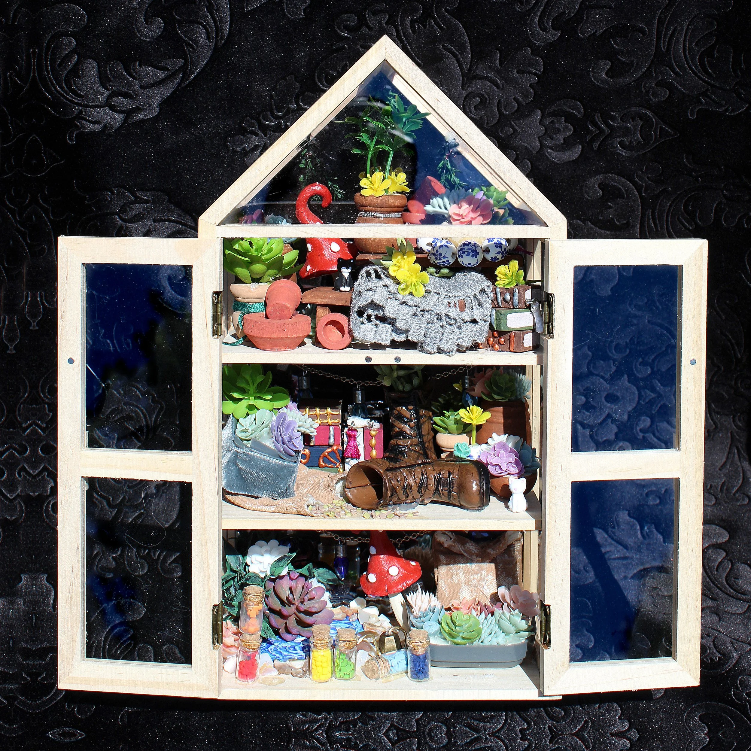 A miniature greenhouse model filled with small plants, succulents, decorative objects, and tiny bottles, set against a dark textured background.