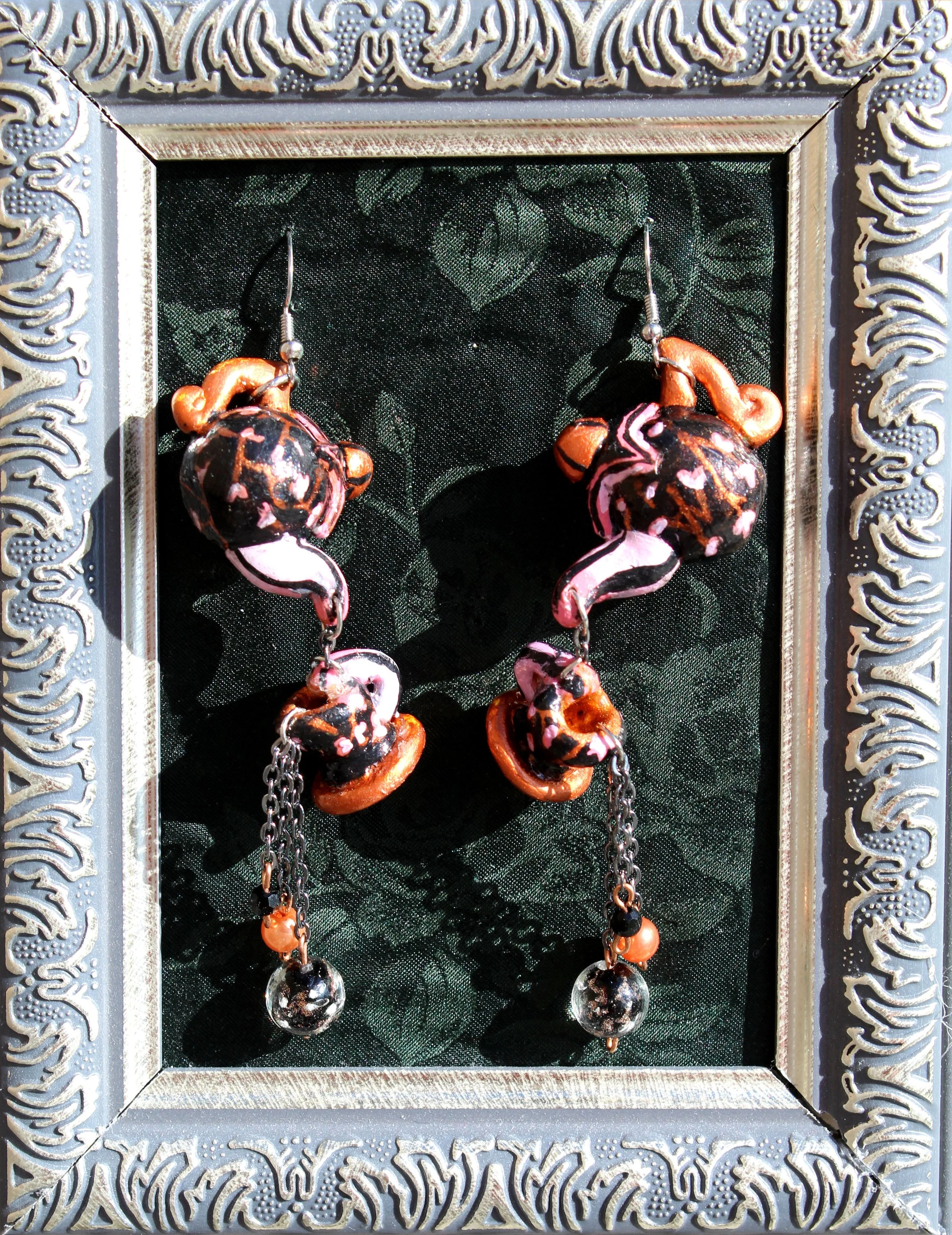 Colorful earrings with black, white, orange, and pink patterns hanging on a decorative black and silver background within a silver ornate picture frame.